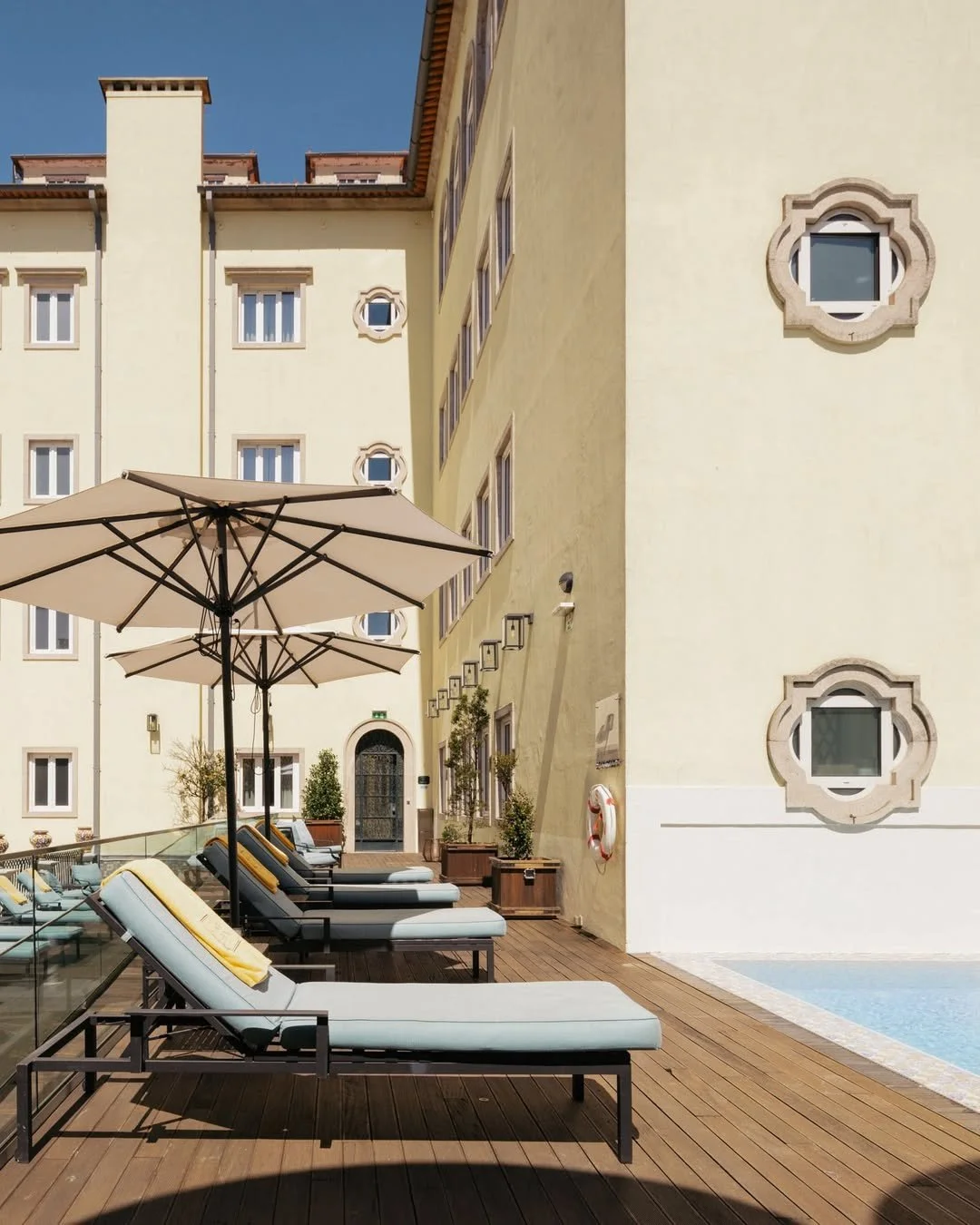 Poolside area with lounge chairs, large umbrella, potted plants, and beige building with decorative window frames.
