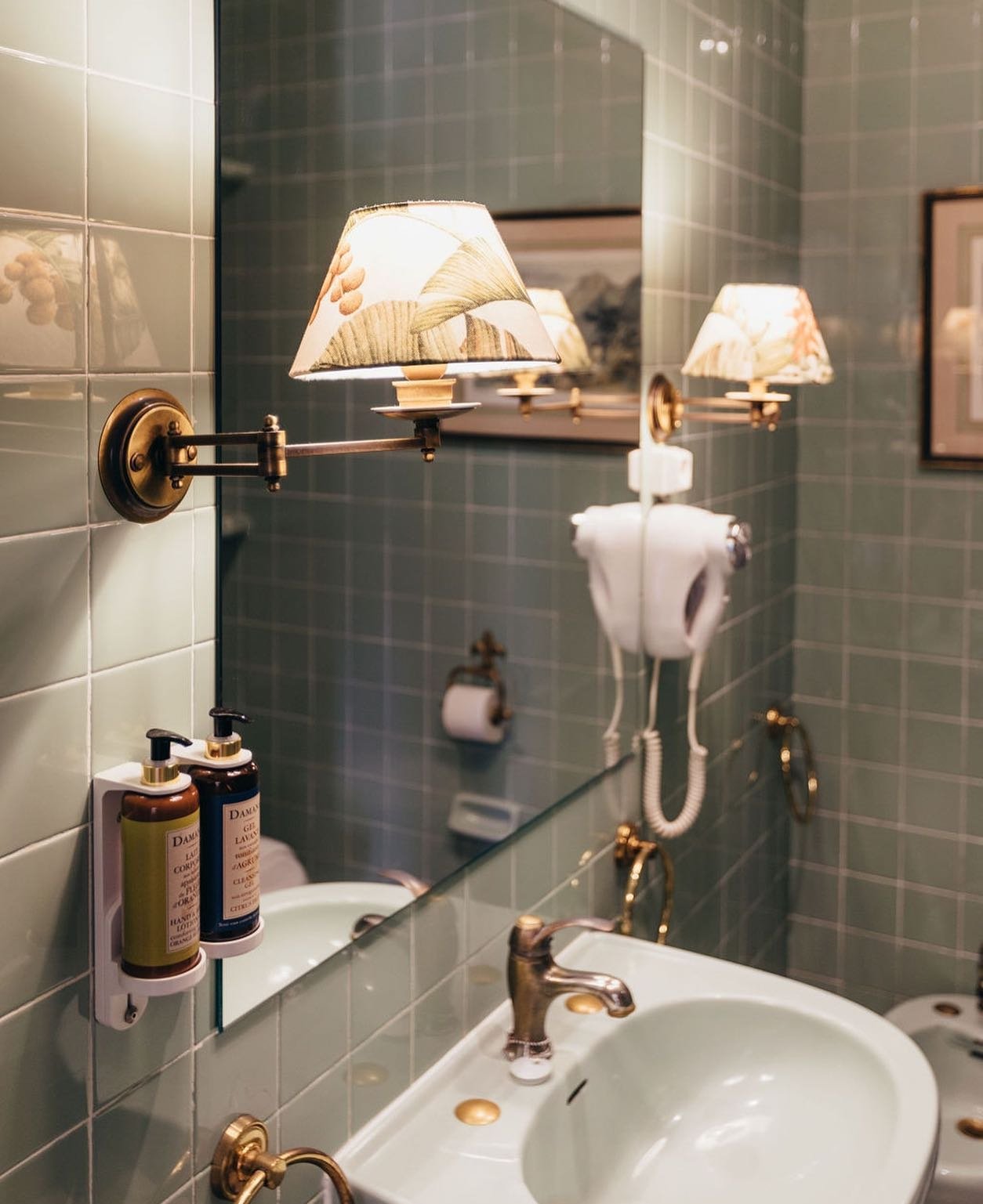 A bathroom with green tiled walls, a mirror, a wall-mounted vintage-style brass lamp with a floral shade, a white sink with a gold faucet, bottles of hand soap, a wall-mounted hairdryer, and a picture frame on the wall.