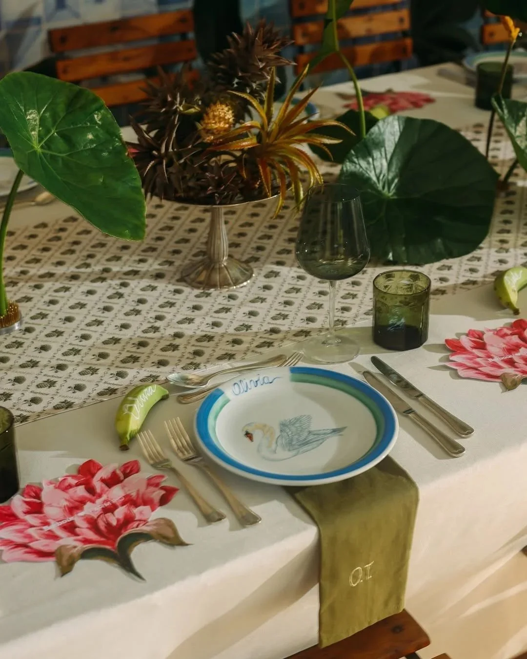 A decorated dining table with floral elements, empty wine glass, dark green drinking glasses, a personalized plate with a swan illustration and the name "Olivia," and utensils arranged for a meal. The table has a green table runner with the initials "O.T." and a floral-themed tablecloth underneath.