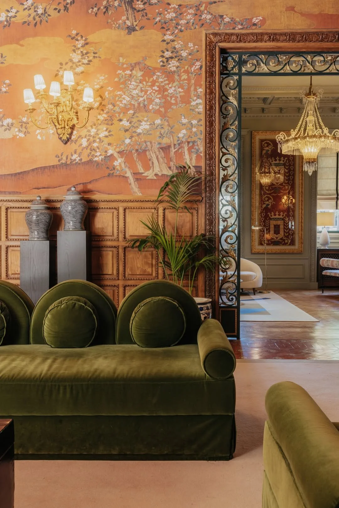 An elegant interior room featuring a green velvet sofa with round pillows, a wooden paneled wall with a mural of trees and a chandelier, potted plant, decorative vases, and ornate metal divider leading to another section with a chandelier and additional furniture.