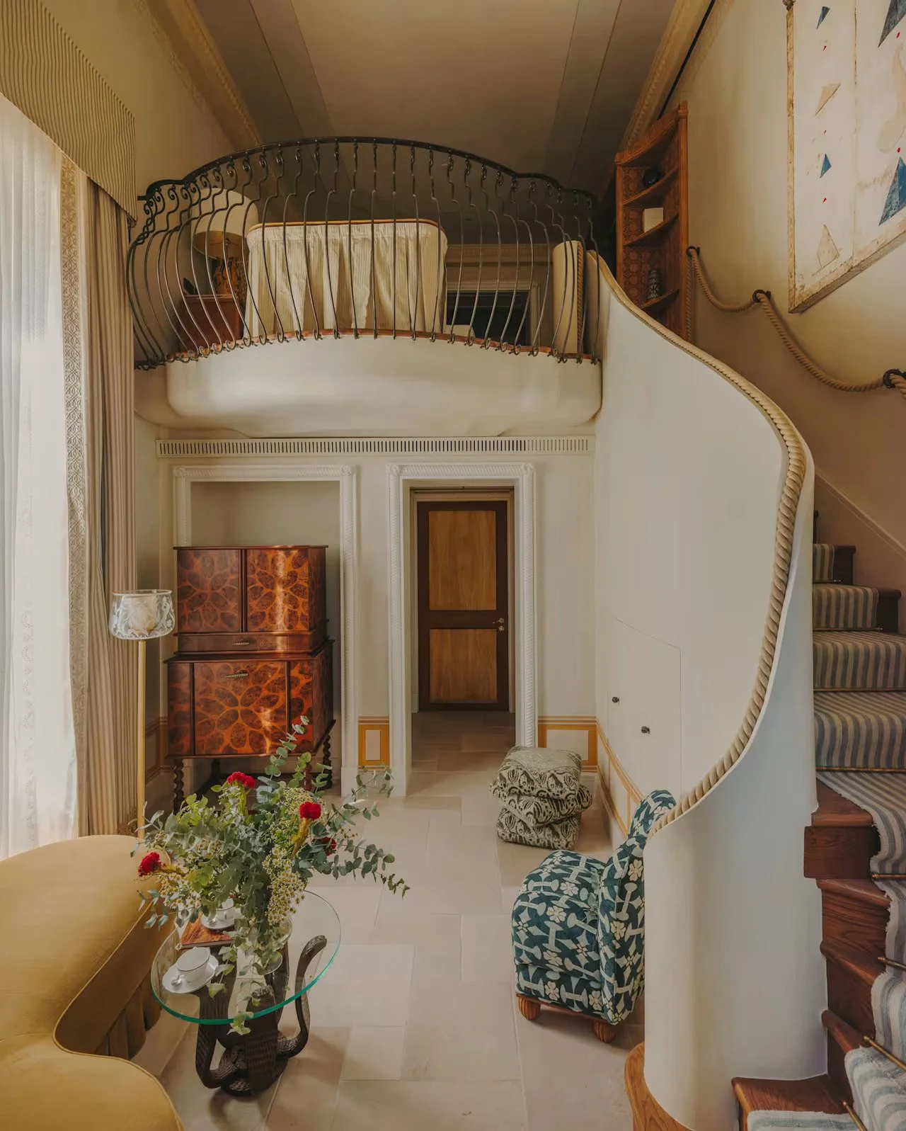 Interior of a multi-level house showing a staircase, a small table with a floral arrangement, a patterned armchair, and a doorway leading to another room. The upper level has a bed with a metal railing and curtains.