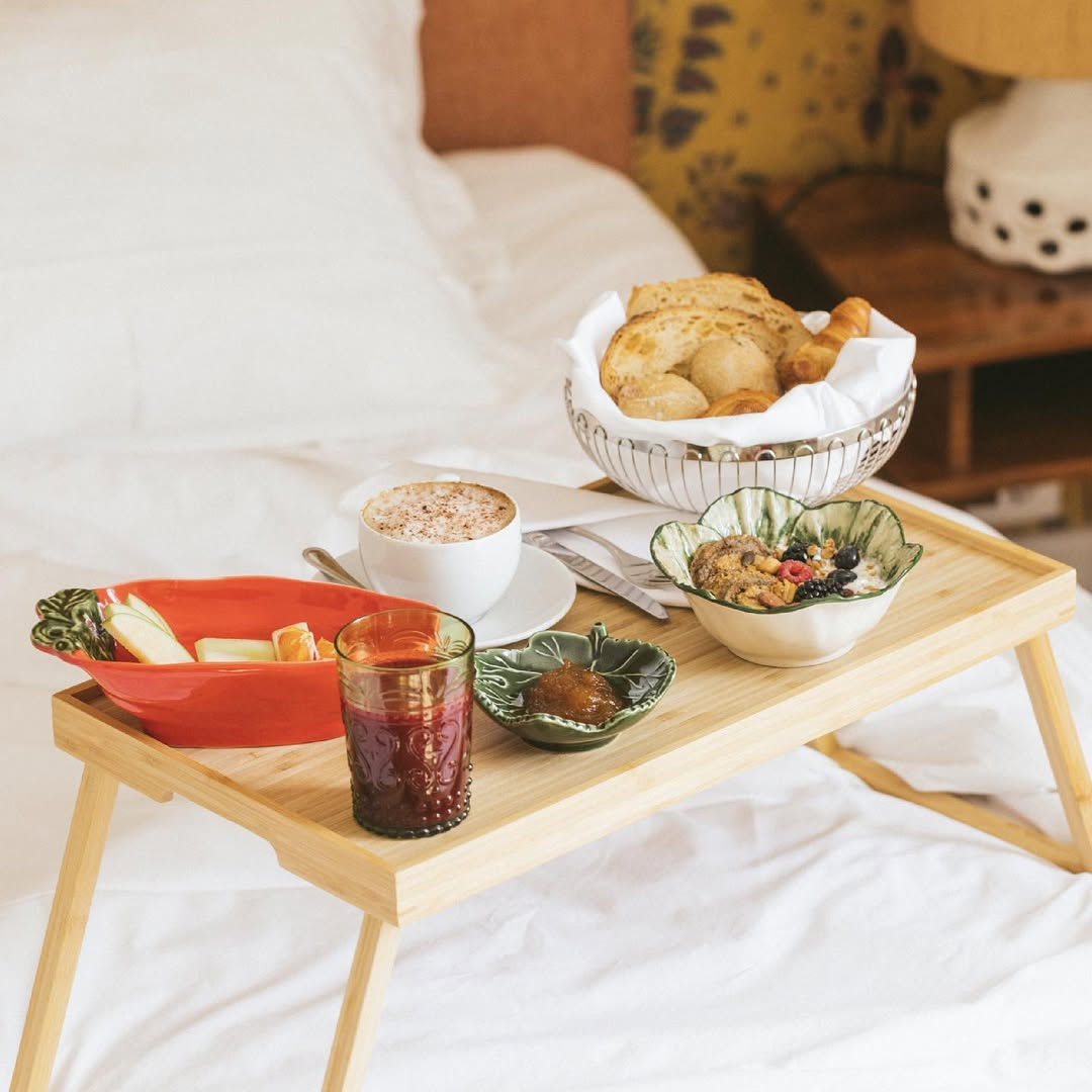 Breakfast tray with various foods on a bed, including bread, fruit, yogurt, cereal, and a drink.