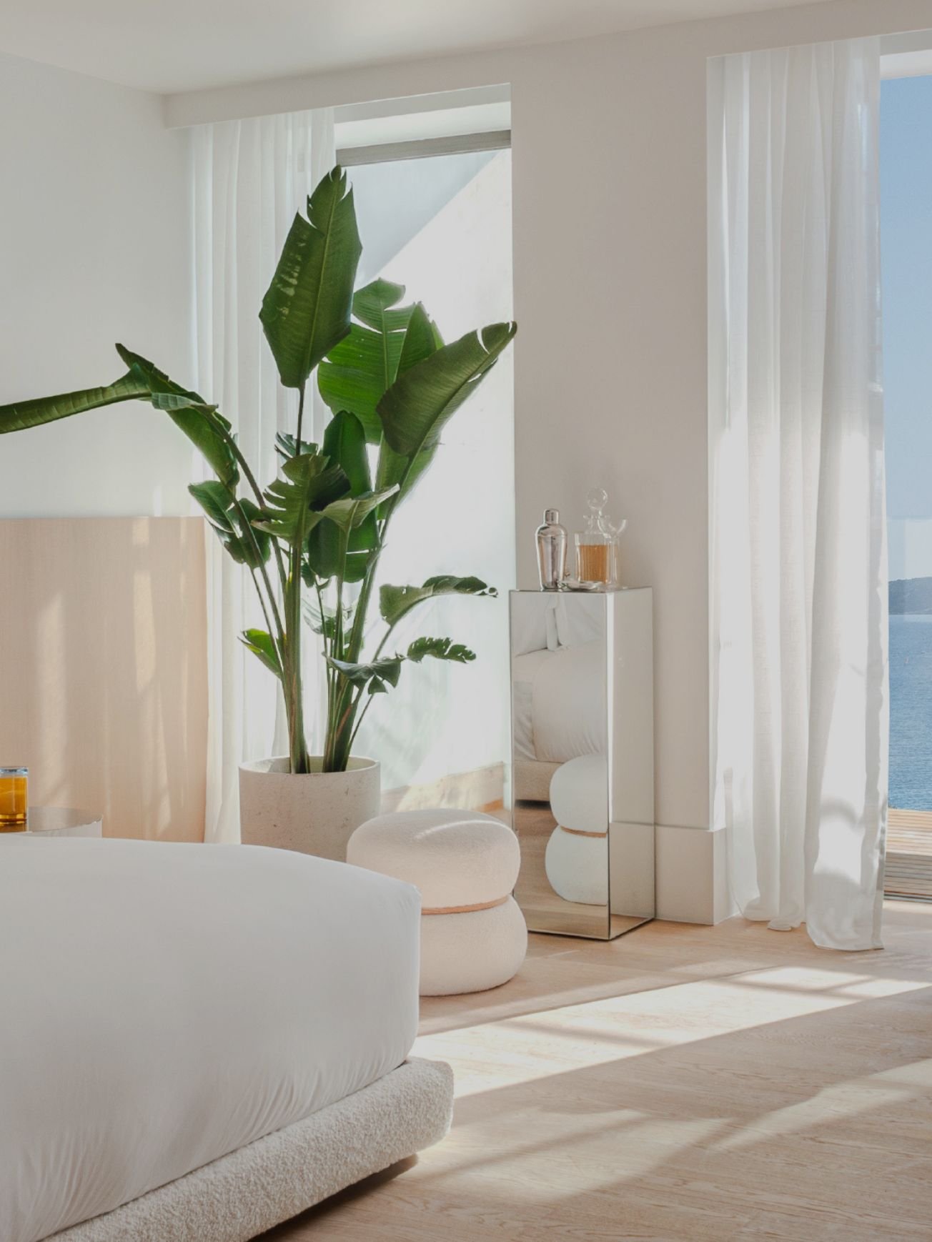 A bright, airy room with large windows covered by sheer white curtains. There is a tall potted plant with large green leaves near the window, and elegant decor including glass bottles on a reflective mirrored cabinet. The room has light-colored wooden flooring, white furniture, and an ocean view visible through the window.