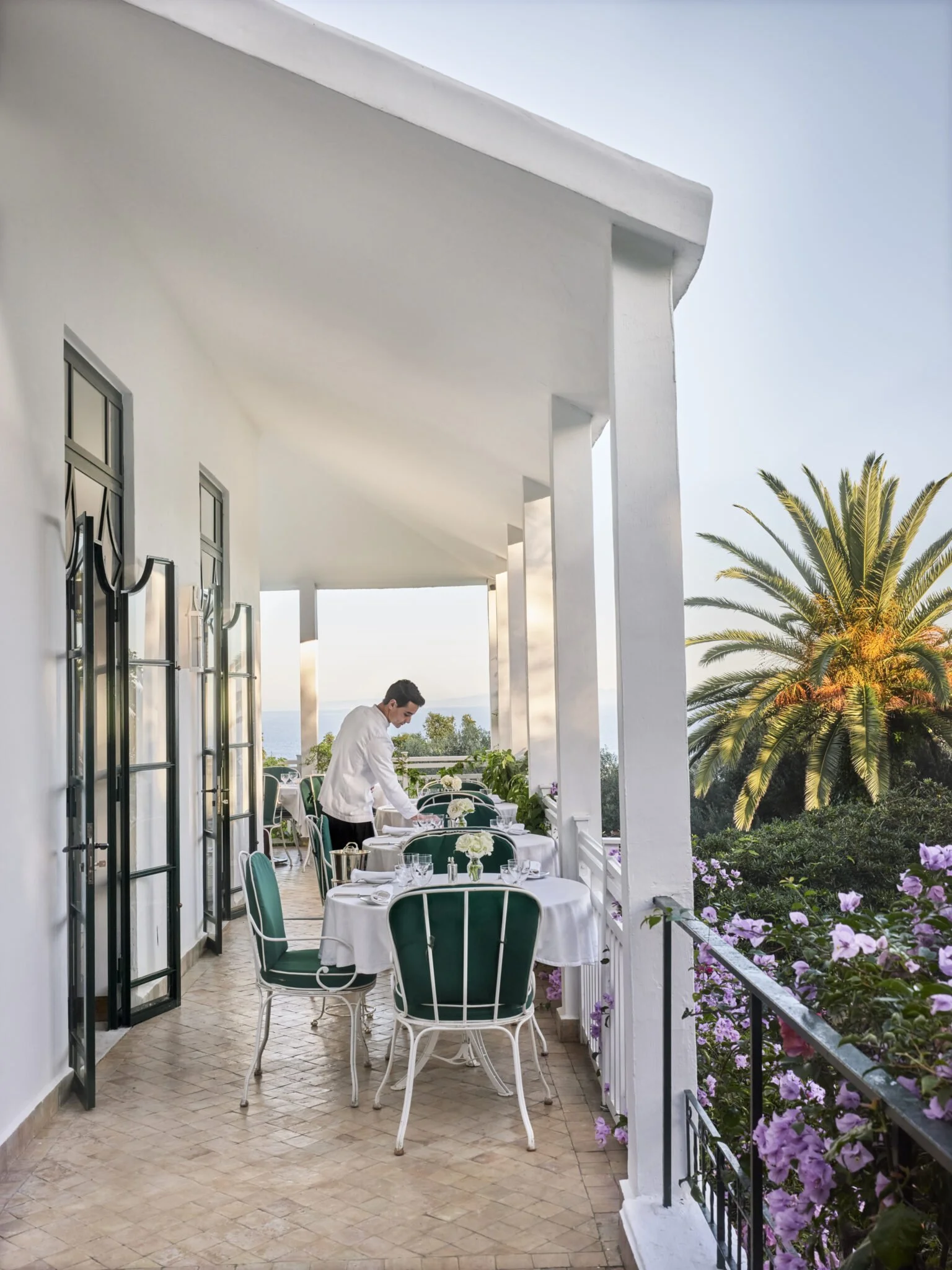 A balcony with white walls and green chairs set for a meal, decorated with flowers, overlooking a scenic view with palm trees and water in the distance, during sunset.