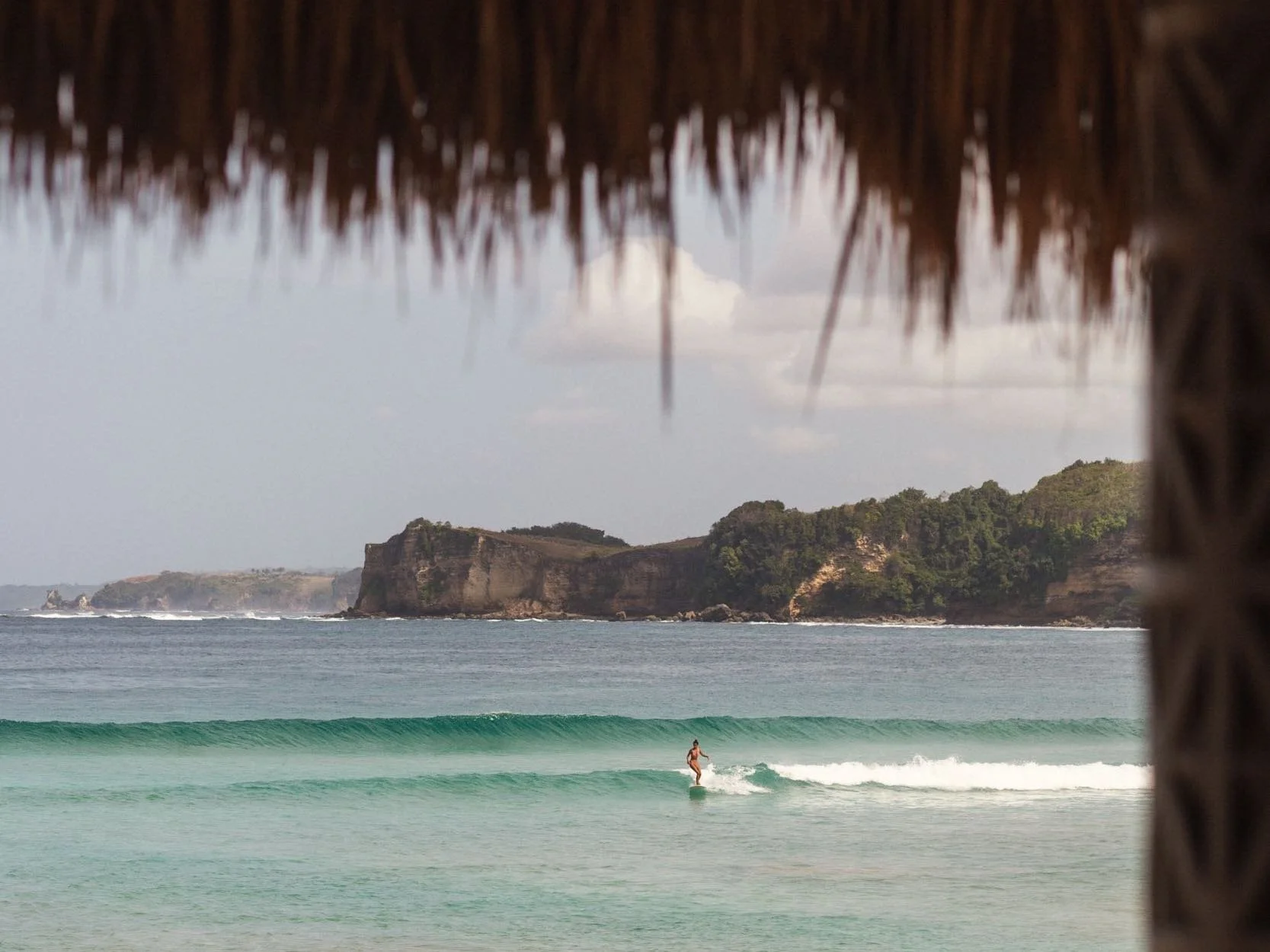 A person surfing in the ocean near a rocky island with green trees, framed by a thatched roof in the foreground.