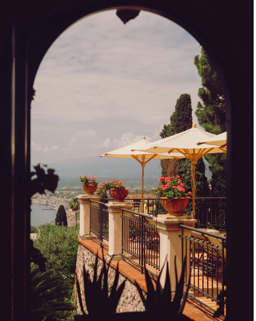 View of a terrace with potted pink flowers, beige umbrellas, and a scenic landscape including trees, a river, and distant mountains, seen through an arched window.
