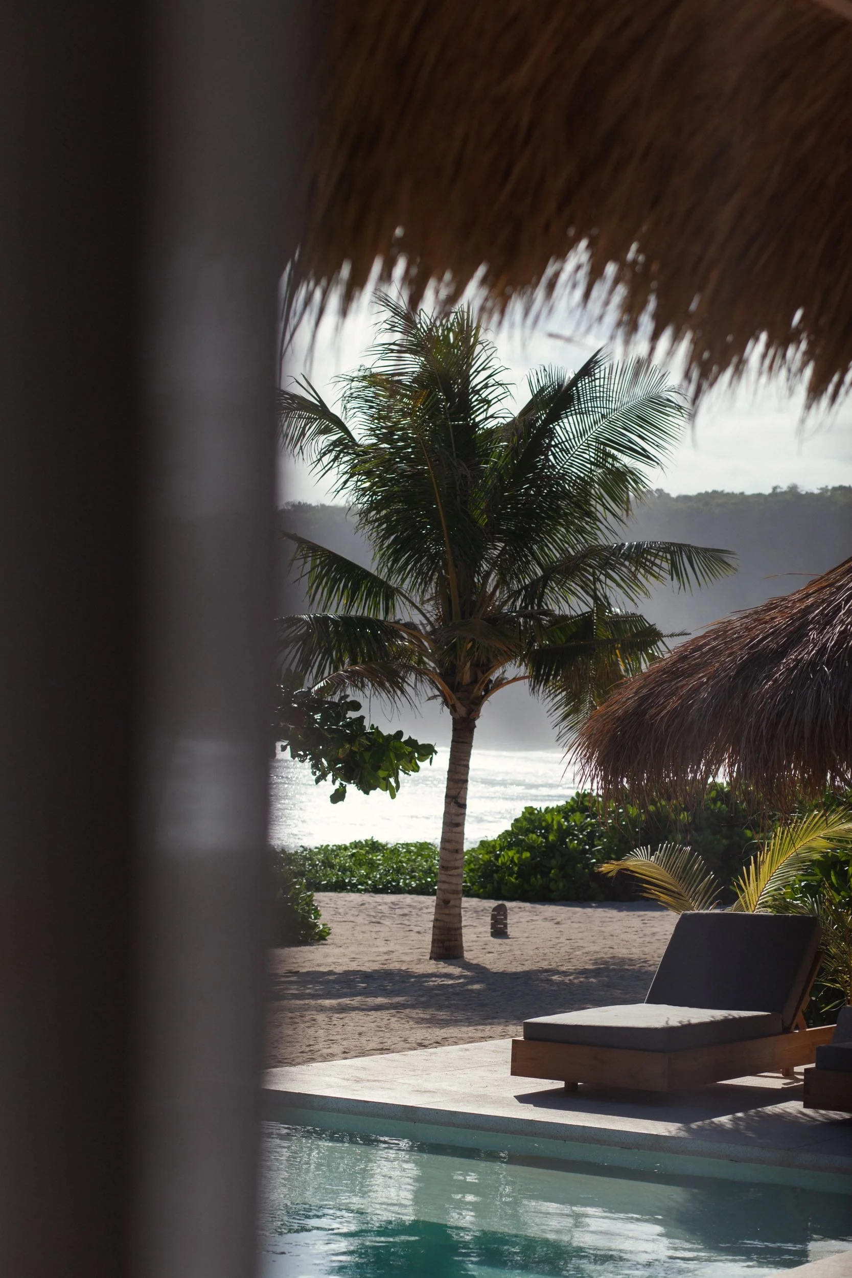 A tropical beach scene with a palm tree, thatched roof structures, and a lounge chair near the water, viewed through a partially enclosed area.