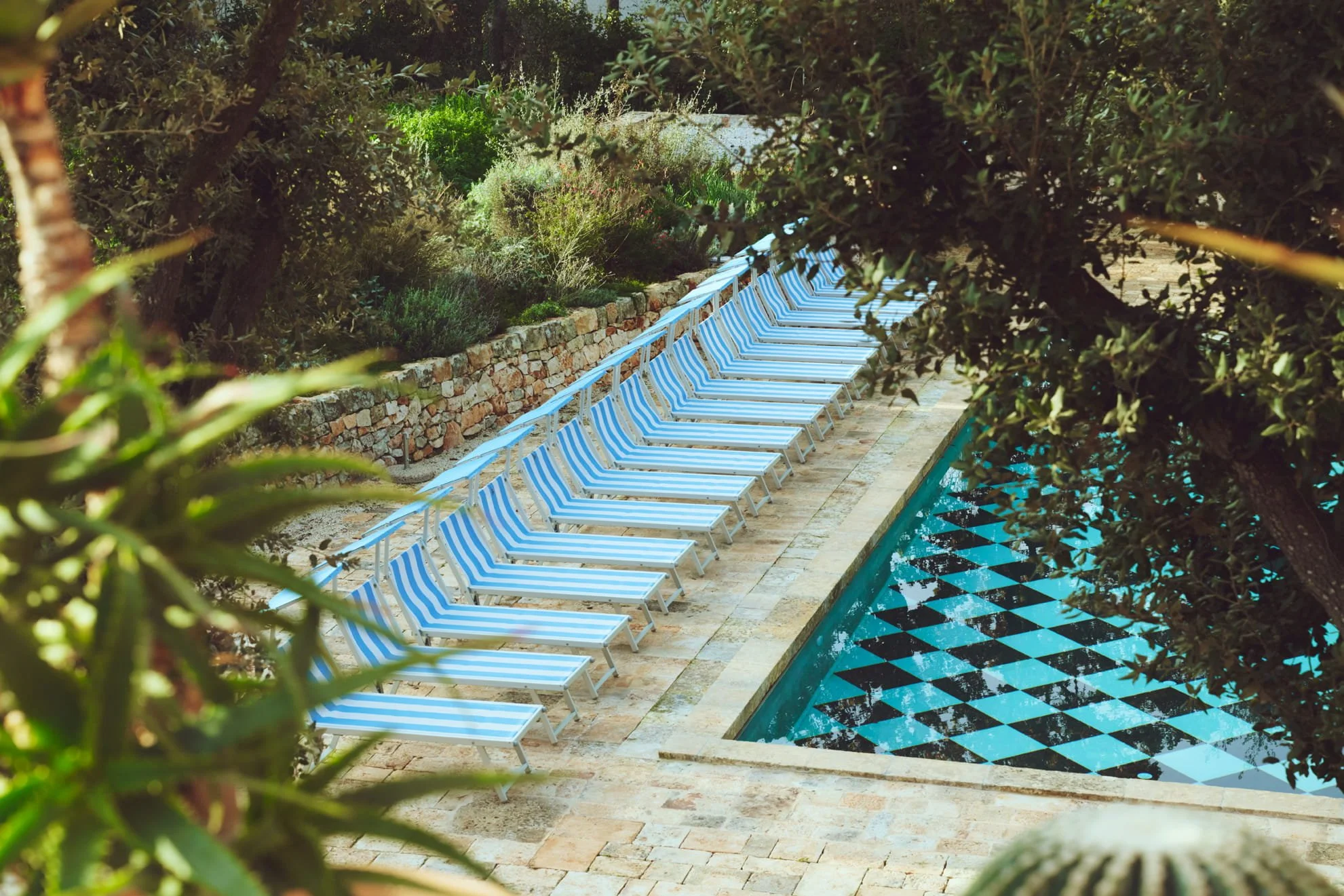 Row of blue and white striped lounge chairs along a swimming pool, surrounded by lush greenery and stone landscaping.