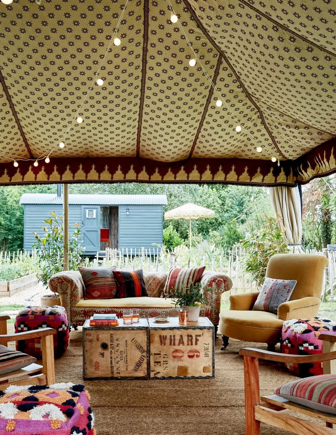 Indoor outdoor seating area under a large decorative tent with string lights, a patterned sofa, armchair, colorful blankets, a wooden table with books and glasses, and a garden view with a small blue shed and patio umbrella.
