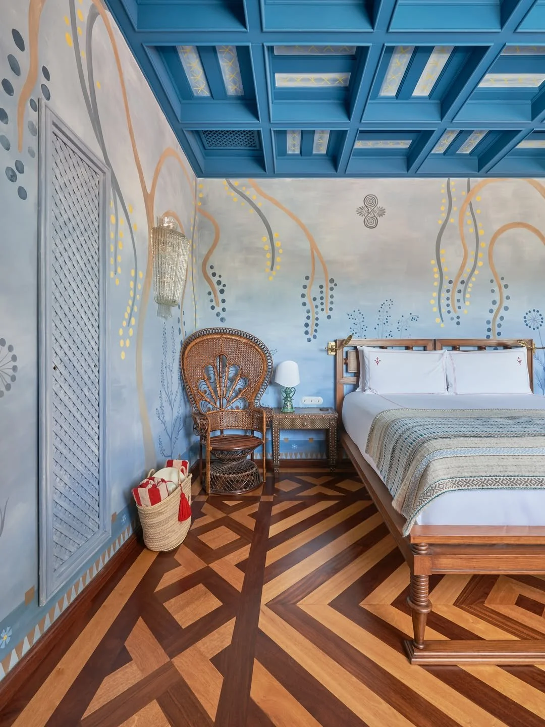 Luxury bedroom with a wooden bed frame, white bedding, and decorative pillows, a wicker chair with a high back, a small side table with a lamp, a basket of rolled towels, a painted wall with colorful abstract patterns, a blue coffered ceiling, and intricate geometric parquet flooring.
