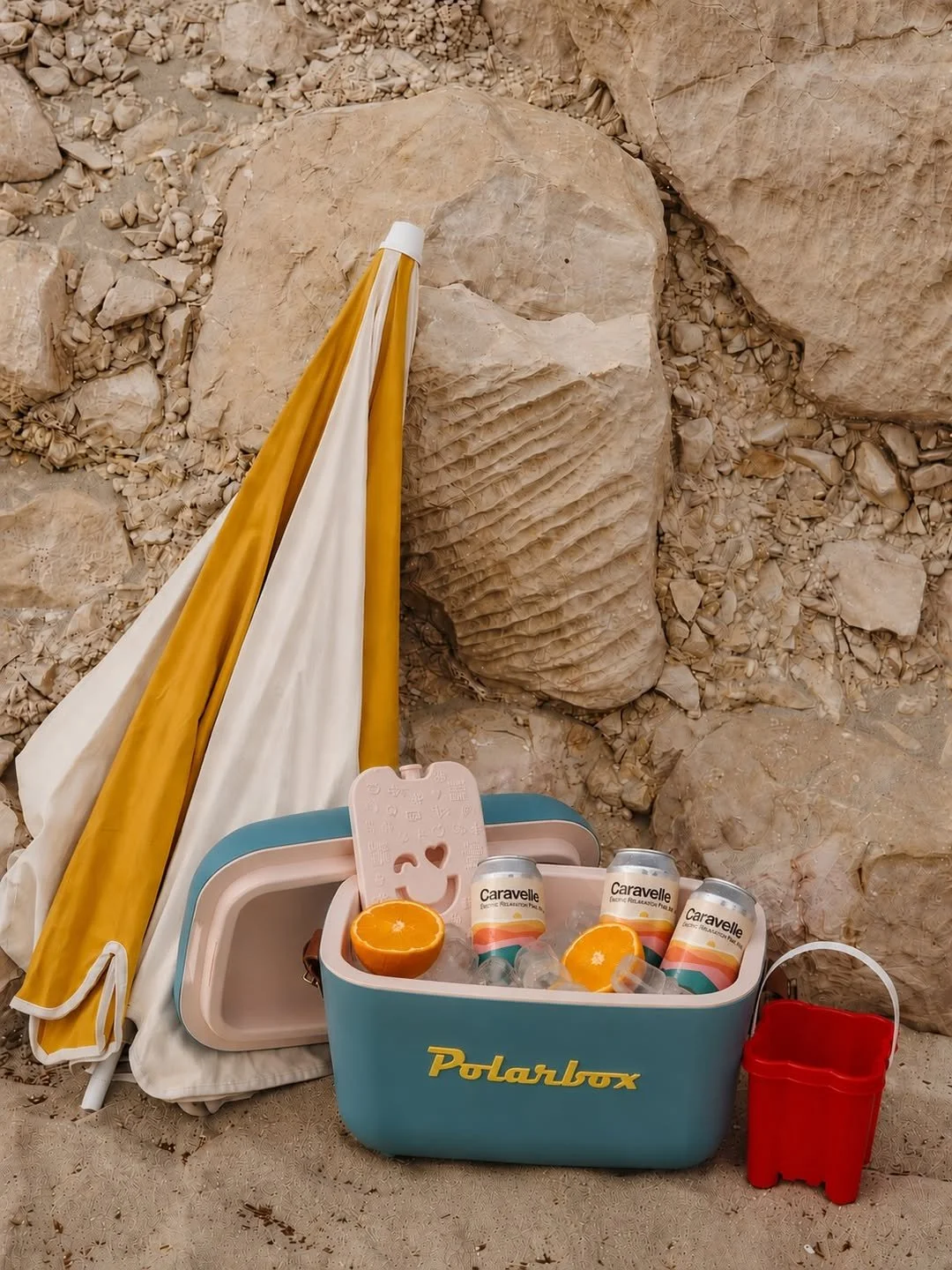A Polarbox cooler filled with cans of Caravelle beverages, halved oranges, and ice cubes, with a pink ice pack, set against a rocky background with a yellow and white beach umbrella leaning on the rocks.