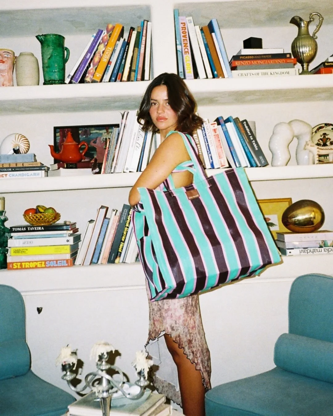 A young woman with shoulder-length brown hair stands in a room with white bookshelves filled with books and decorative items. She wears a floral dress with a large striped tote bag over her shoulder, and stands between two blue armchairs, with a silver candelabra in front of her.