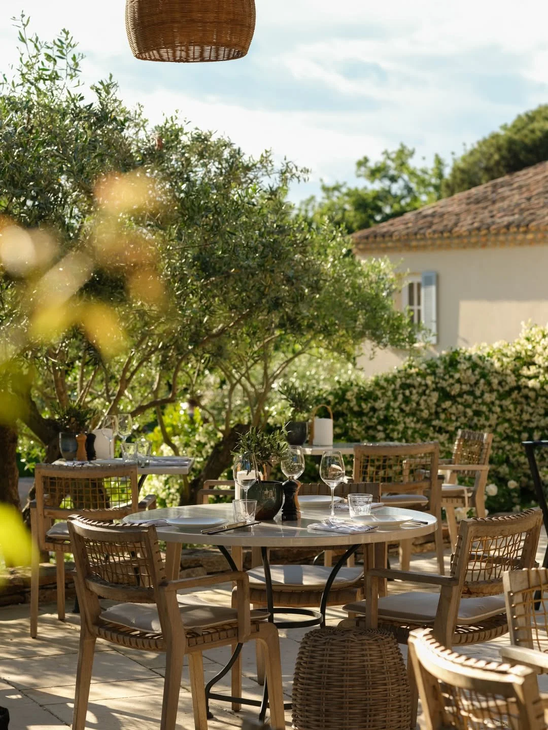Outdoor dining area with a round table set for four, surrounded by wooden chairs with white cushions, under trees with green foliage, with a house in the background. Table includes wine glasses, a pepper grinder, and decorative plants.