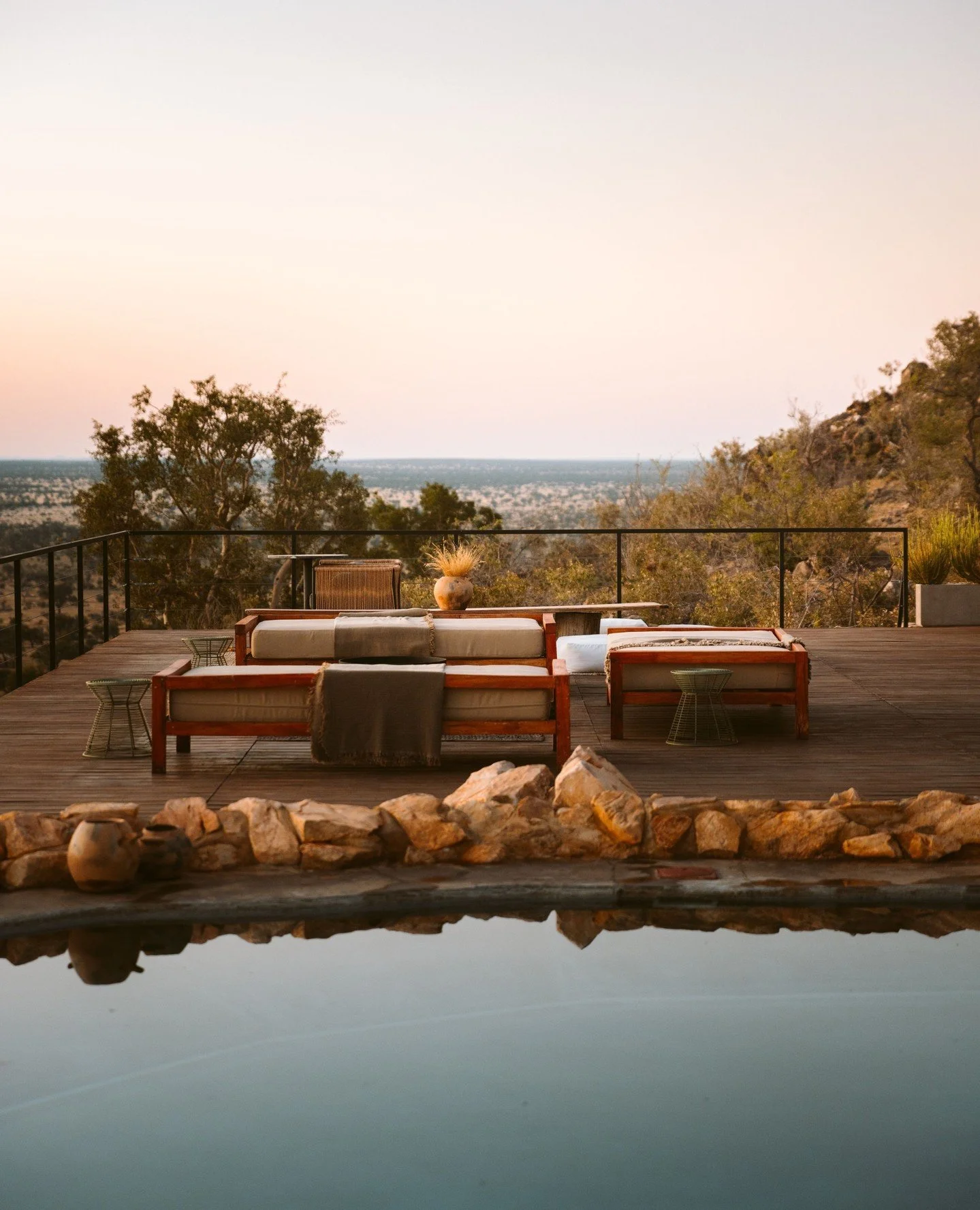An outdoor patio with wooden flooring, modern seating, and decorative elements, overlooking a scenic landscape with trees and a hazy horizon at sunset.