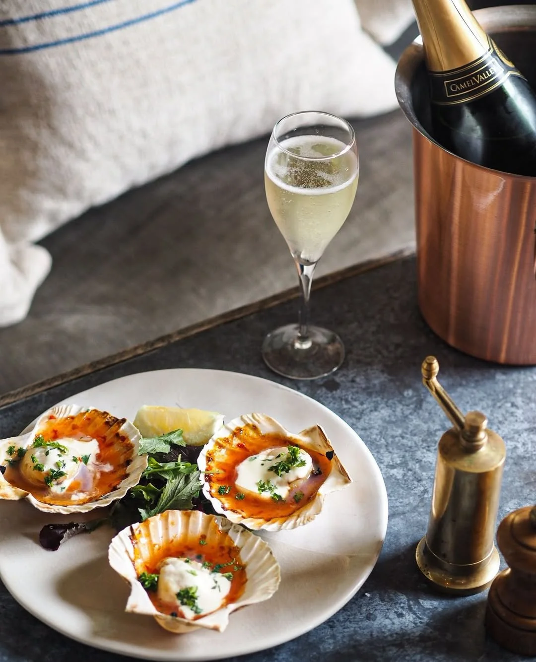 A plate of baked oysters garnished with herbs, a wedge of lemon, a glass of champagne, and an ice bucket with a bottle of champagne.