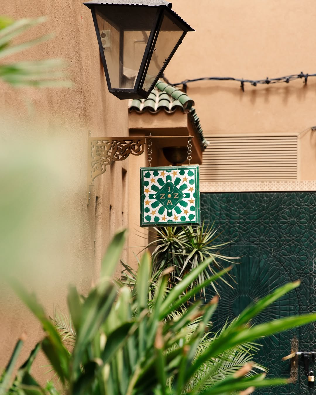 Decorative sign hanging on a beige wall with lush green plants in the foreground and a lantern overhead.