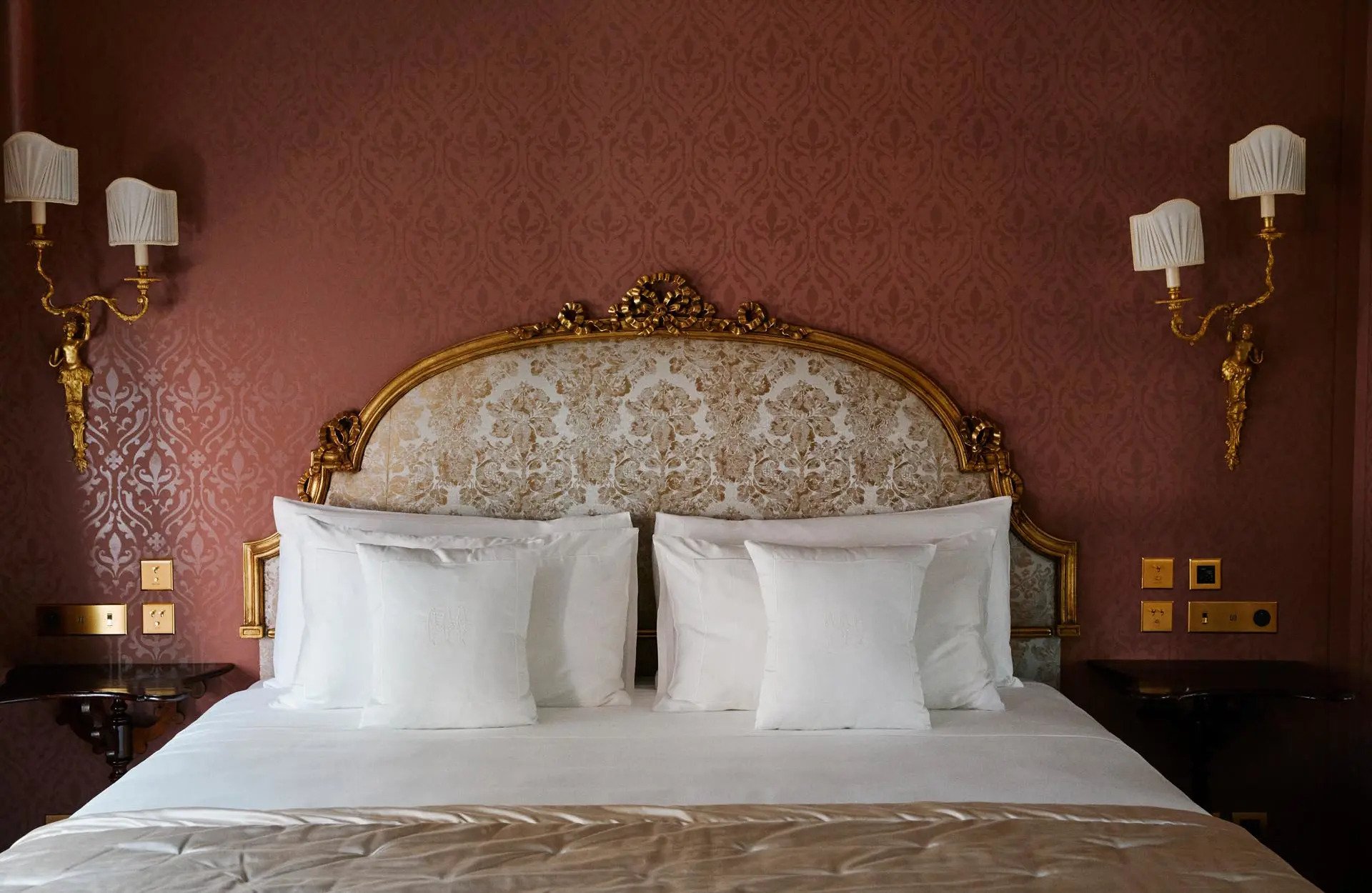 A hotel bed with white bedding and pillows, ornate gold headboard, pink patterned wall with gold wall sconces on either side, and light switches visible on the wall.