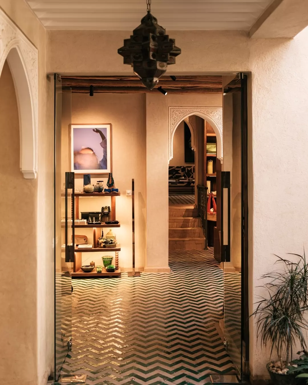 Interior view of a room with Moroccan-style decor, featuring a patterned tile floor, a wall with built-in shelves displaying decorative items, and an archway leading to another room with stairs. There is a hanging lantern and a potted plant by the doorway.