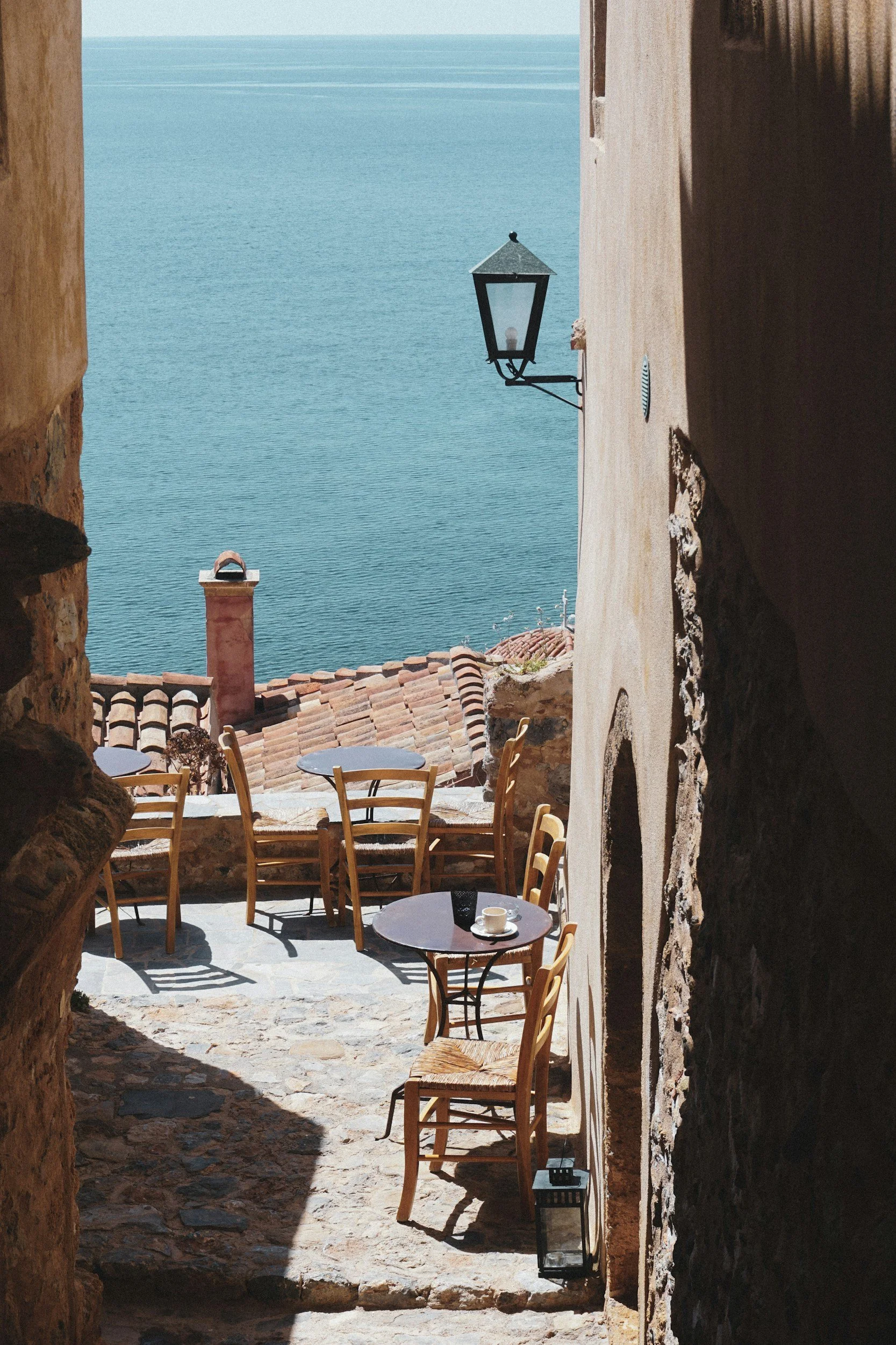 A cozy terrace with round tables and chairs, view of the sea, and a tiled roof, viewed through a stone archway.