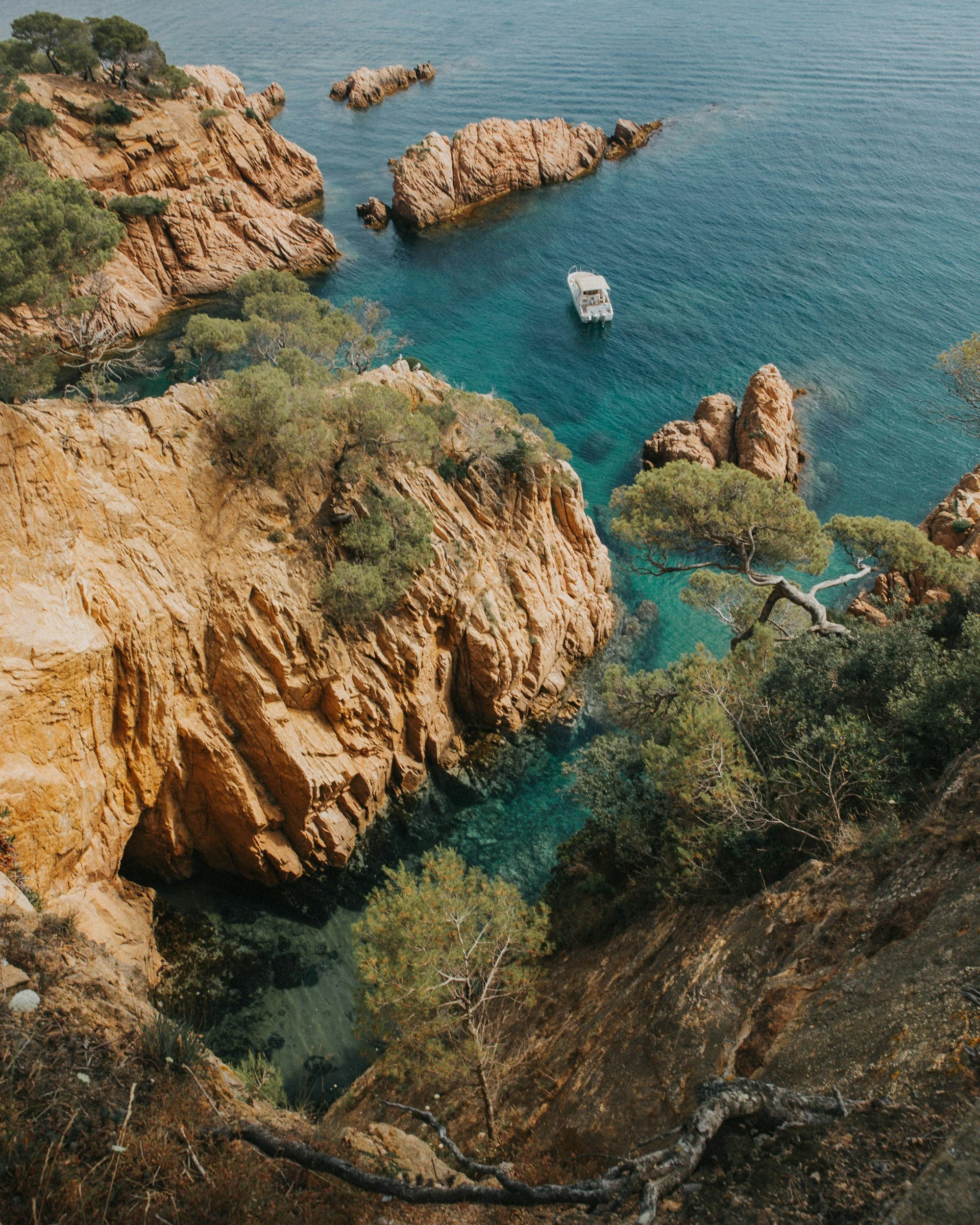 Bird's eye view of a rocky coastline with turquoise water, a small white boat, and lush green trees on cliffs.
