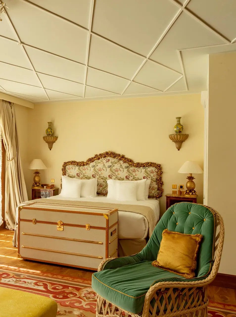 A luxurious hotel room decorated in vintage style with a large bed having an ornate, floral-patterned headboard, a cloth trunk at the foot of the bed, a green carved wicker armchair with a gold pillow, and matching lamps on bedside tables. The room has cream-colored walls, a decorative ceiling, and a gold-colored rug.