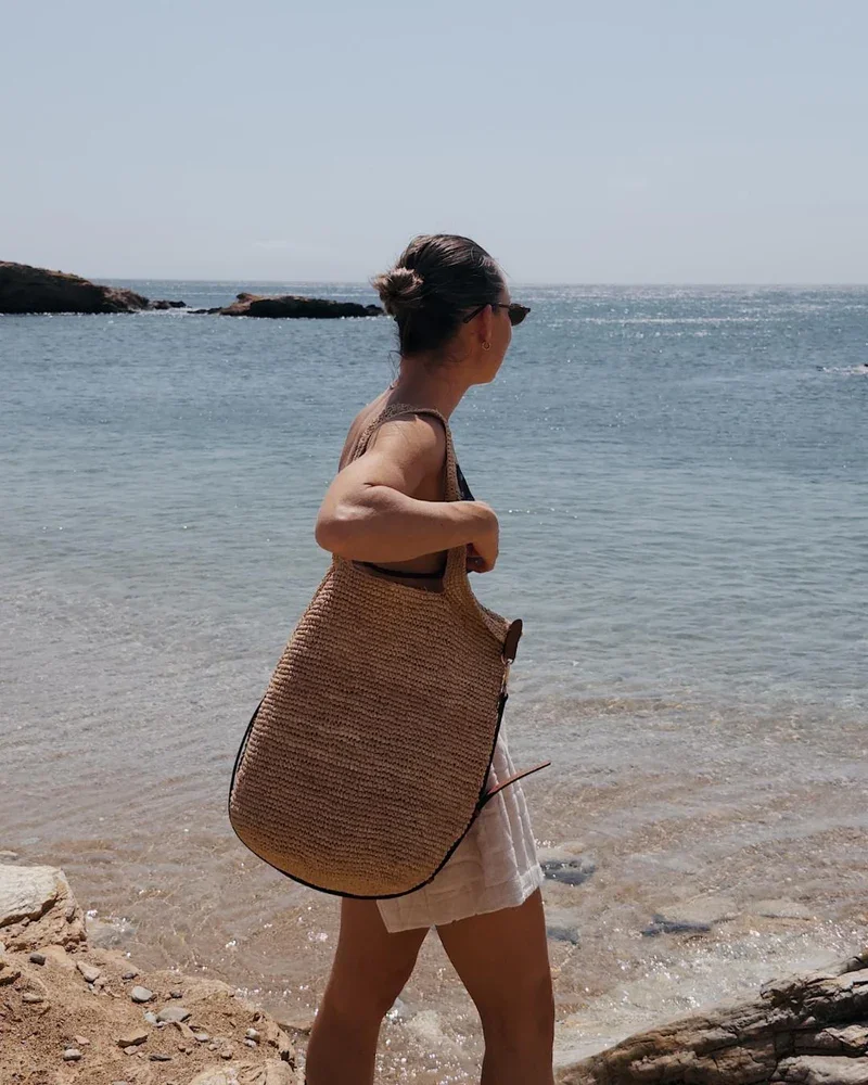 A woman walking along a beach with a large woven bag, wearing sunglasses, a sleeveless top, and shorts, with the ocean and rocks in the background.