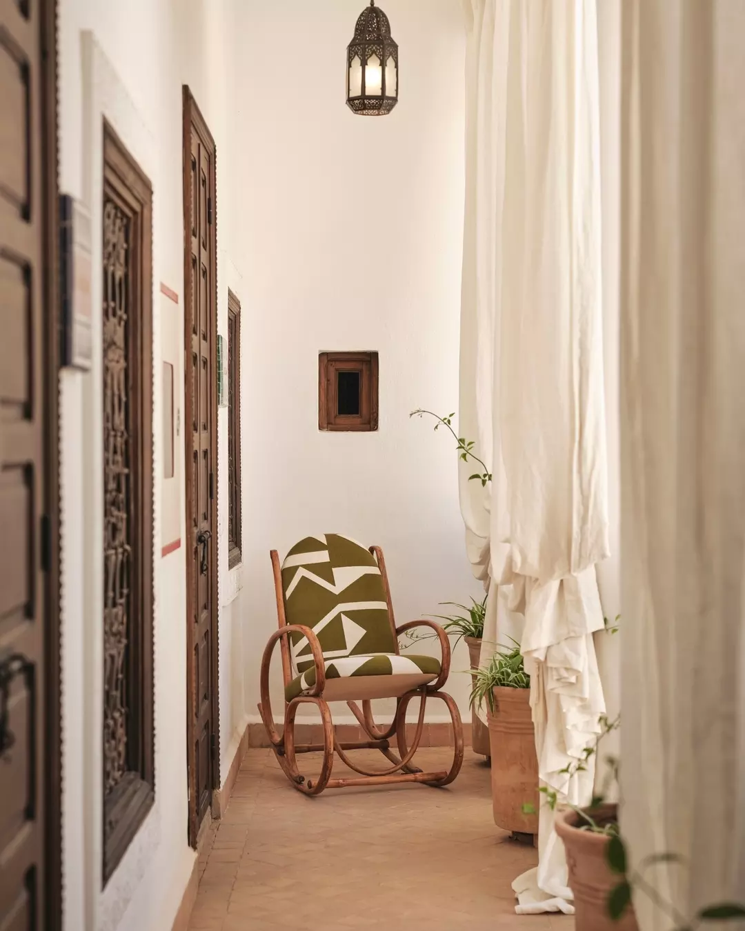 A cozy porch with a wooden rocking chair upholstered in green and white fabric, surrounded by potted plants and framed by white curtains. A Moroccan-style lantern hangs from the ceiling.