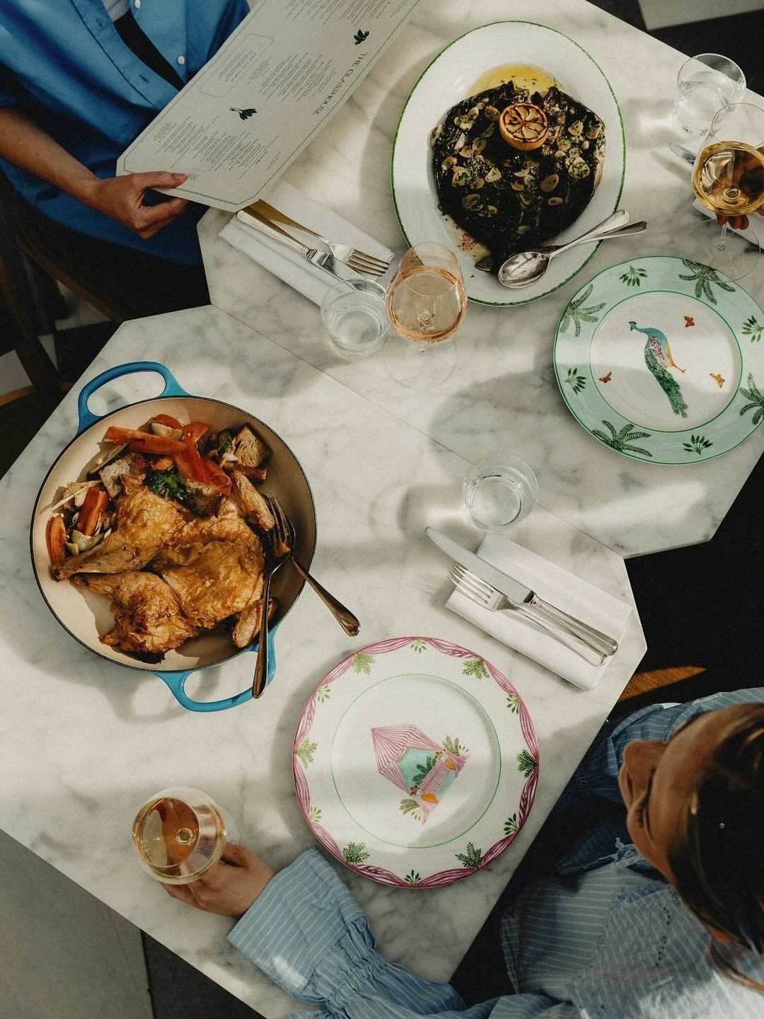 Overhead view of a marble dining table with a roasted chicken dish with vegetables, a chocolate dessert with walnuts and a lemon slice, colorful plates with a peacock and a pink border, and three glasses of rosé wine. Two people are seated at the table, one holding a glass and the other reading a menu.