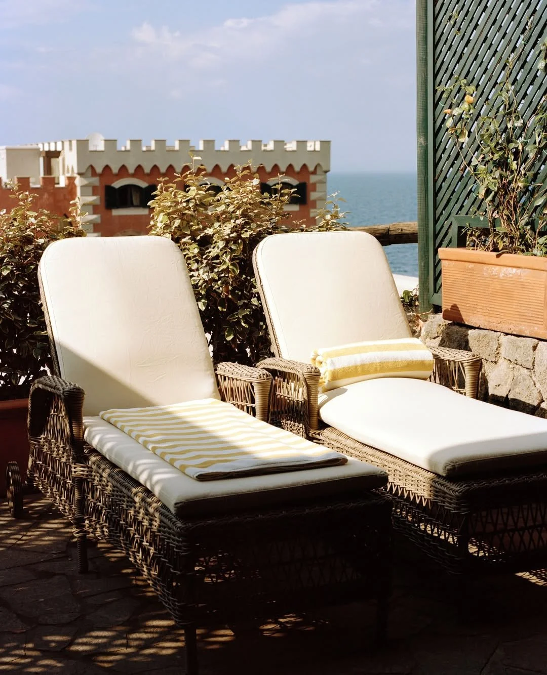 Two wicker lounge chairs with white cushions and yellow and white striped towels on a balcony overlooking the sea and a building with a castle-like rooftop.