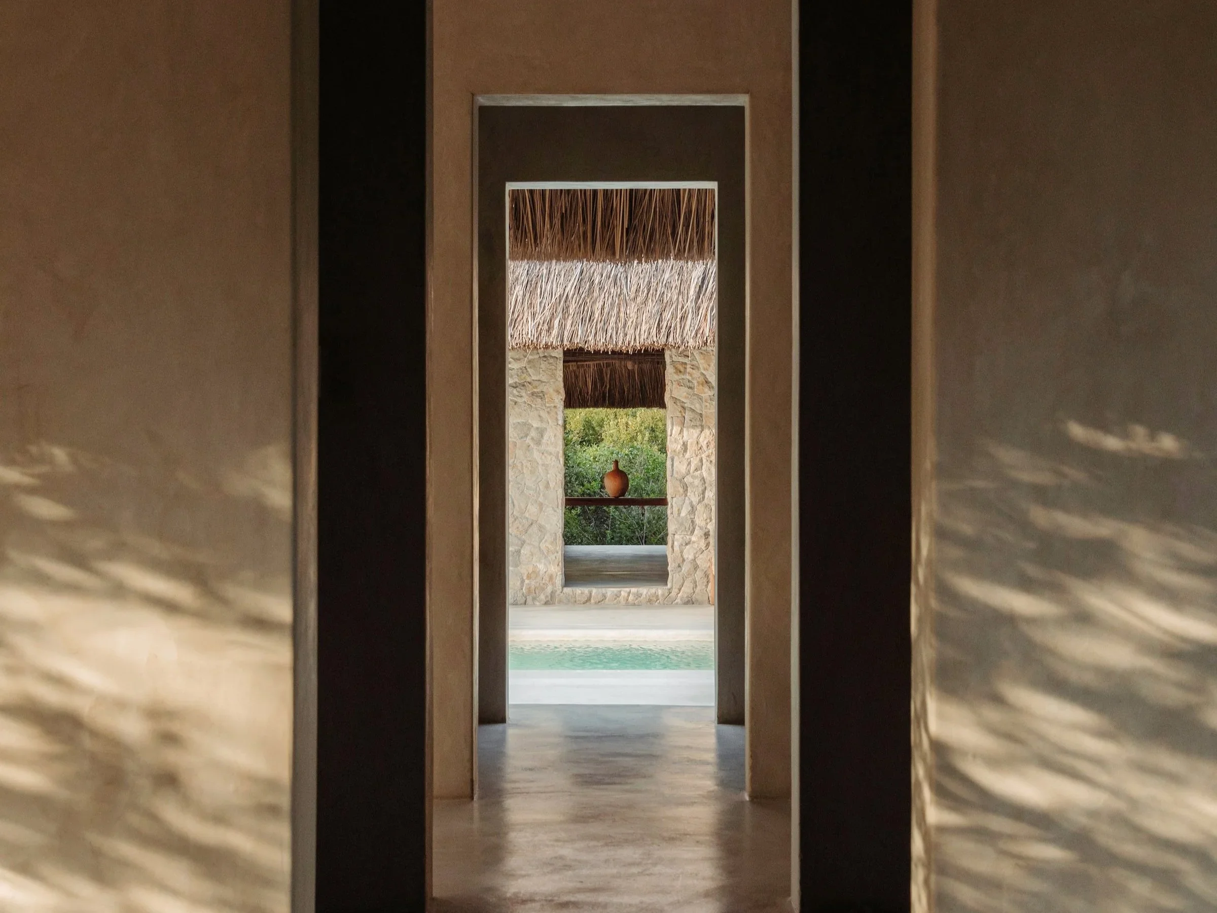 View through a series of doorways leading to an outdoor area with thatched roofing, stone walls, a small decorative statue, and greenery with a pool in the foreground.