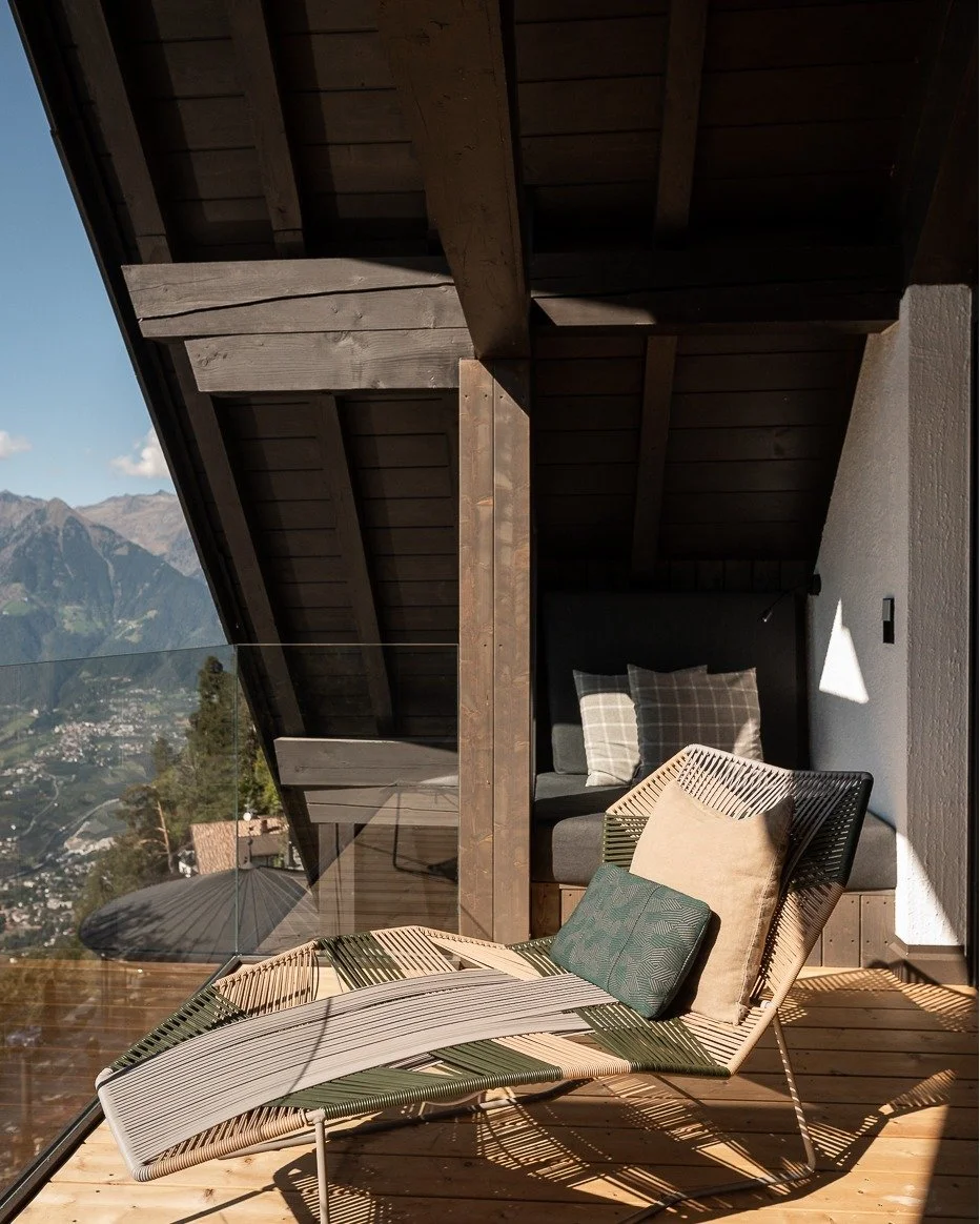 A wooden outdoor balcony with a modern chaise lounge, decorative pillows, a cushioned bench, and a mountain view. The balcony has wooden beams, a glass railing, and is partially shaded.