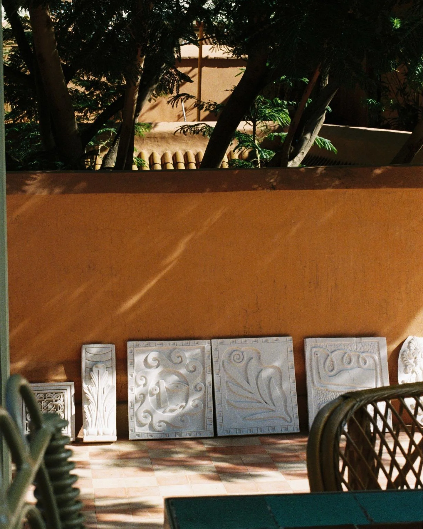 Display of carved stone or plaster panels with ornate patterns, arranged against a wall in a shaded outdoor area, with wicker furniture and potted plants visible.