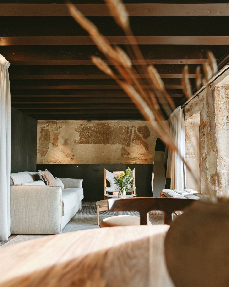 Living room with a cream sofa, wooden chairs, a flower vase on the table, stone walls, a window with white curtains, and a ceiling with wooden beams.