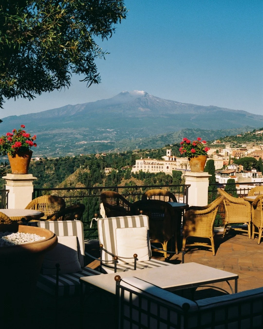 Terrace with outdoor seating and pink flowering plants, overlooking a cityscape with historical buildings, and Mount Vesuvius in the background under a clear blue sky.