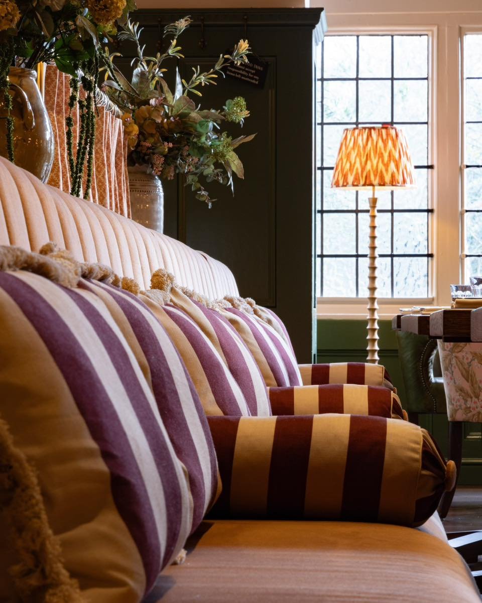 A cozy living room corner with a beige tufted sofa decorated with striped pillows in purple and yellow, a tall floor lamp with a patterned orange lampshade, a large window with grid panes, and a side table with decorative items and a floral tablecloth. There are also large vases with floral arrangements.