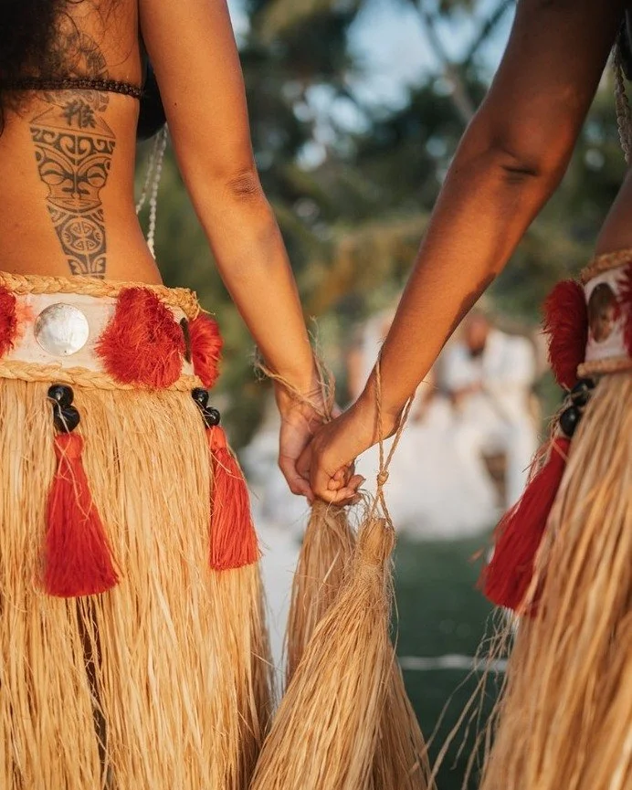 Two people holding hands, wearing traditional grass skirts and celebrating outdoors.