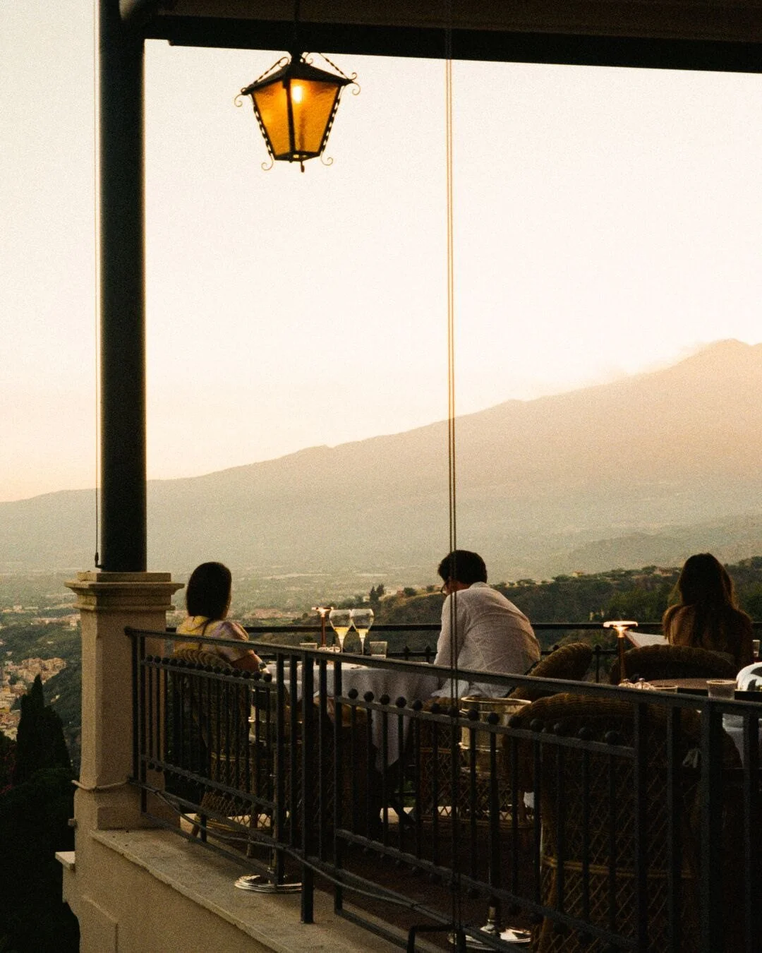 People dining on a balcony with a view of mountains during sunset, with a hanging lantern overhead.
