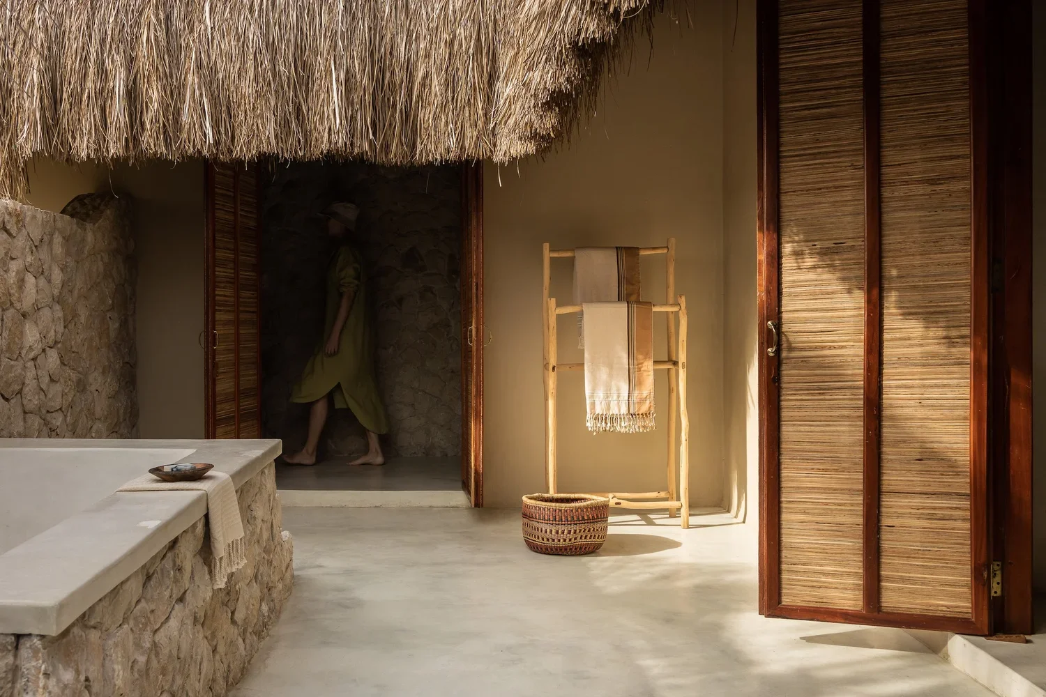 Interior of a room with stone and wooden walls, thatched roof, a wooden towel rack with towels, a wicker basket, and a separate shower area with a wall of stone and a person standing inside.