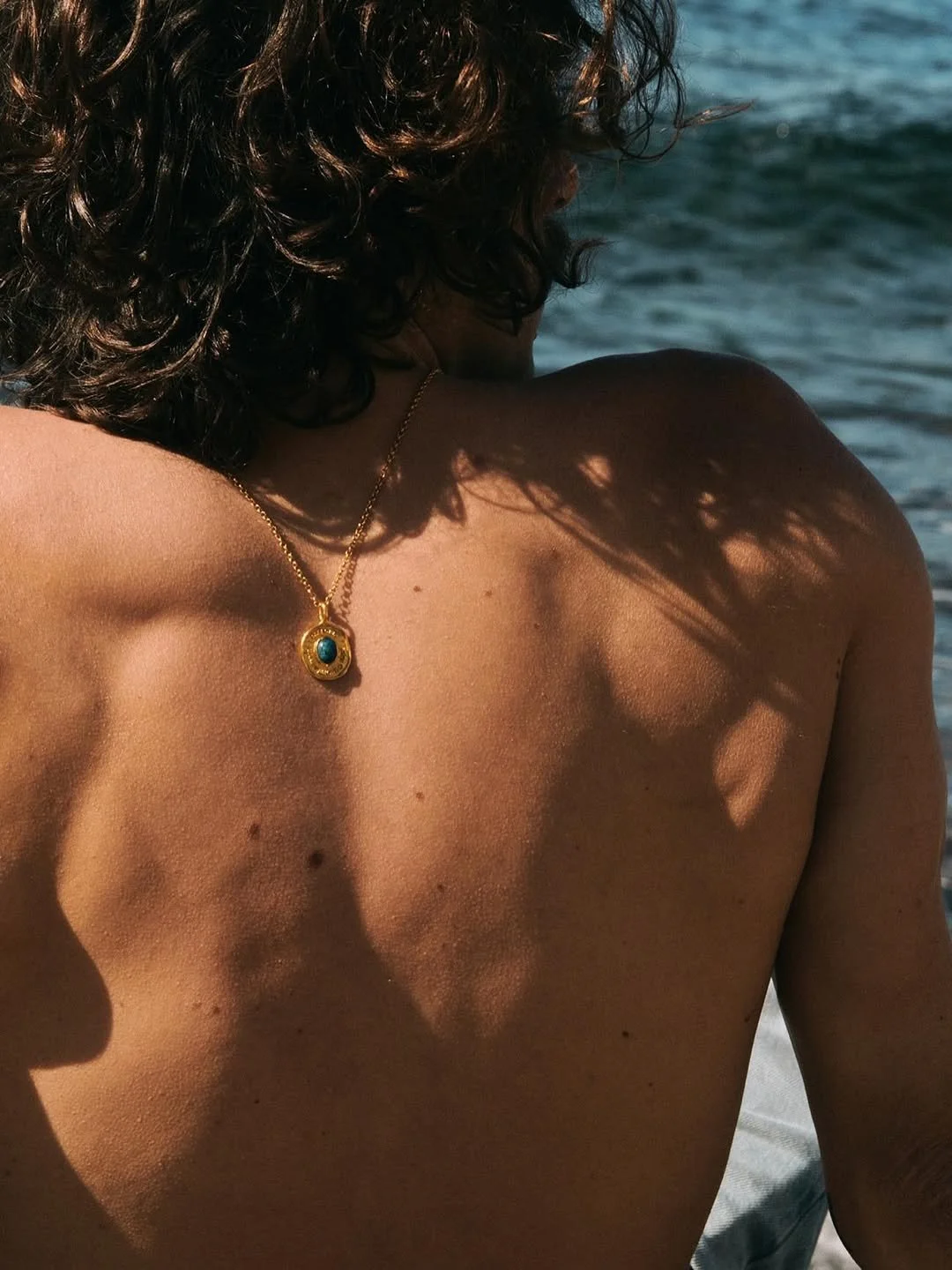 Close-up of a woman's bare back and shoulder, with sunlight casting shadows of palm leaves. She wears a gold necklace with a turquoise pendant. The background shows the ocean.