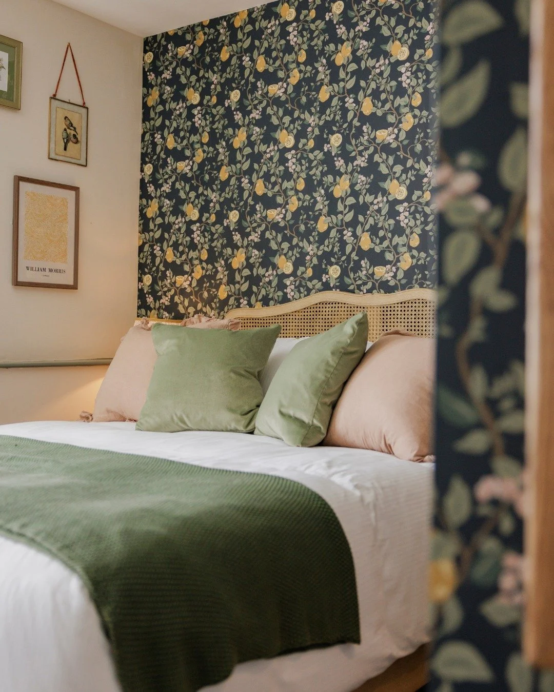 A cozy bedroom with a bed covered in white linens, topped with green and peach pillows, and a green throw blanket. The headboard is woven rattan, and the background wall features dark floral wallpaper with yellow and white flowers. To the left, there are framed pictures on the wall.