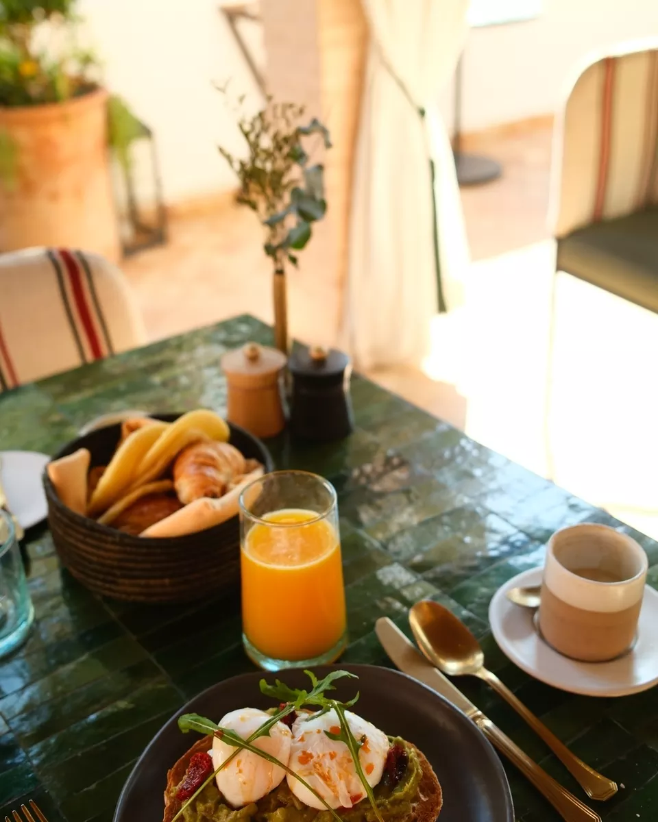 Breakfast table with an orange juice, a cup of coffee, a basket of bread and croissants, and a plate with eggs, avocado, and toppings.