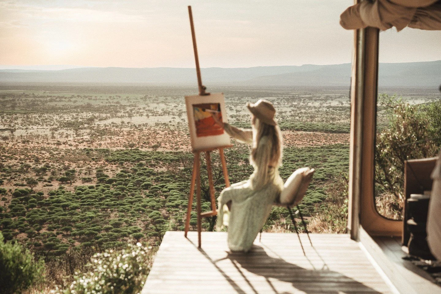 A person sitting on an outdoor chair, painting on an easel, with a vast desert landscape in the background, featuring sparse vegetation and distant mountains.