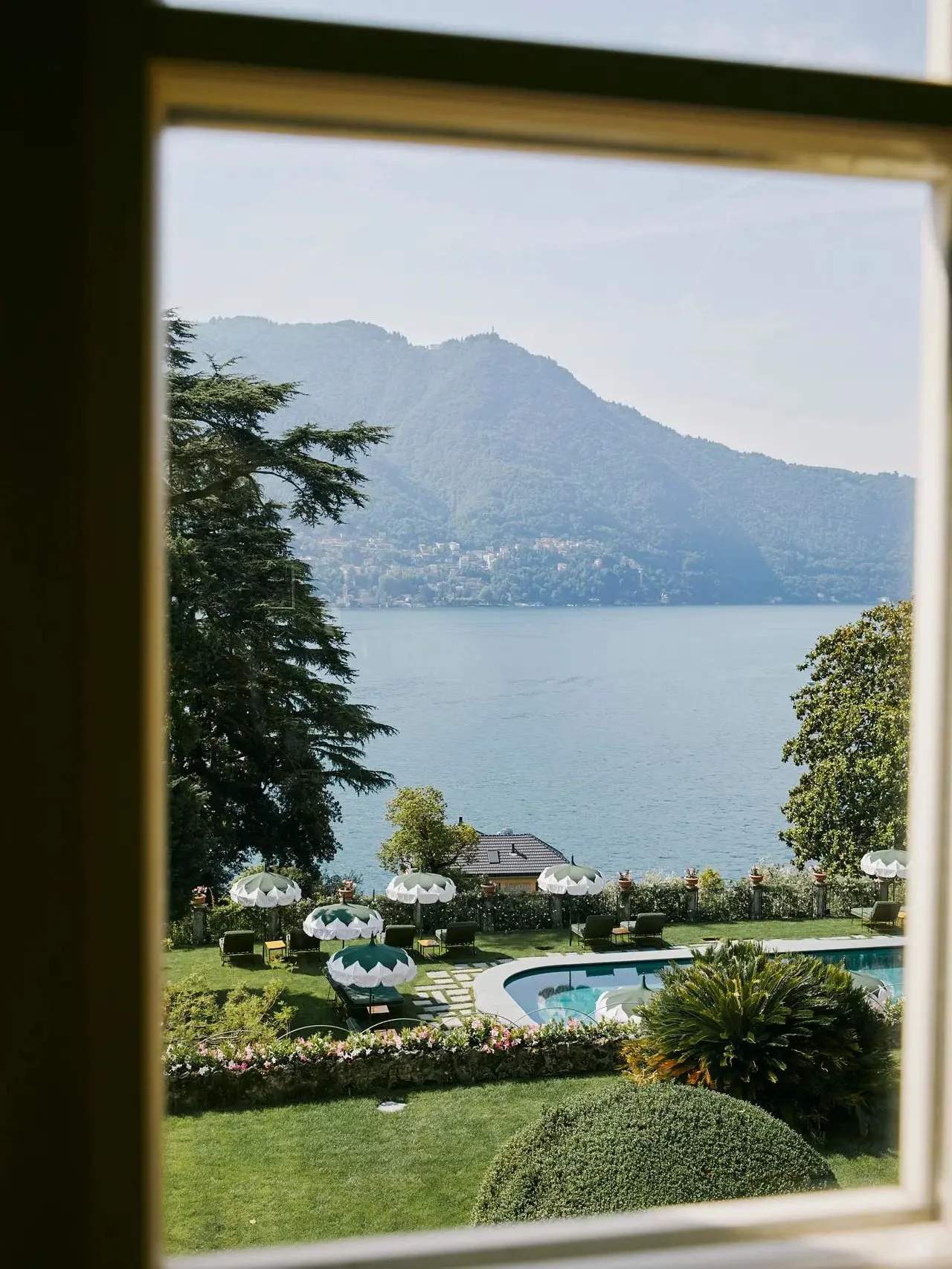View through a window shows a lakeside landscape with a swimming pool, white umbrellas, lounge chairs, a garden with trimmed bushes and flowers, calm water, and mountains in the distance.