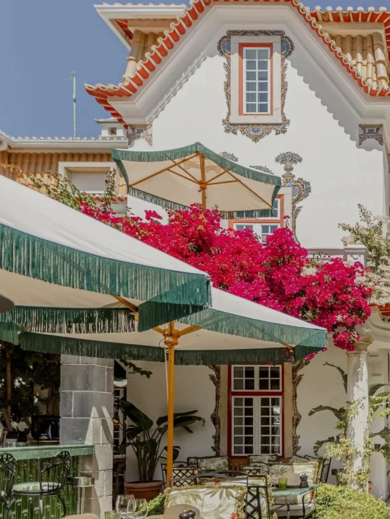 A courtyard with tables and chairs, pink flowering bushes, green and white shade umbrellas, and a decorative house with ornate window frames and a tiled roof, under a clear blue sky.