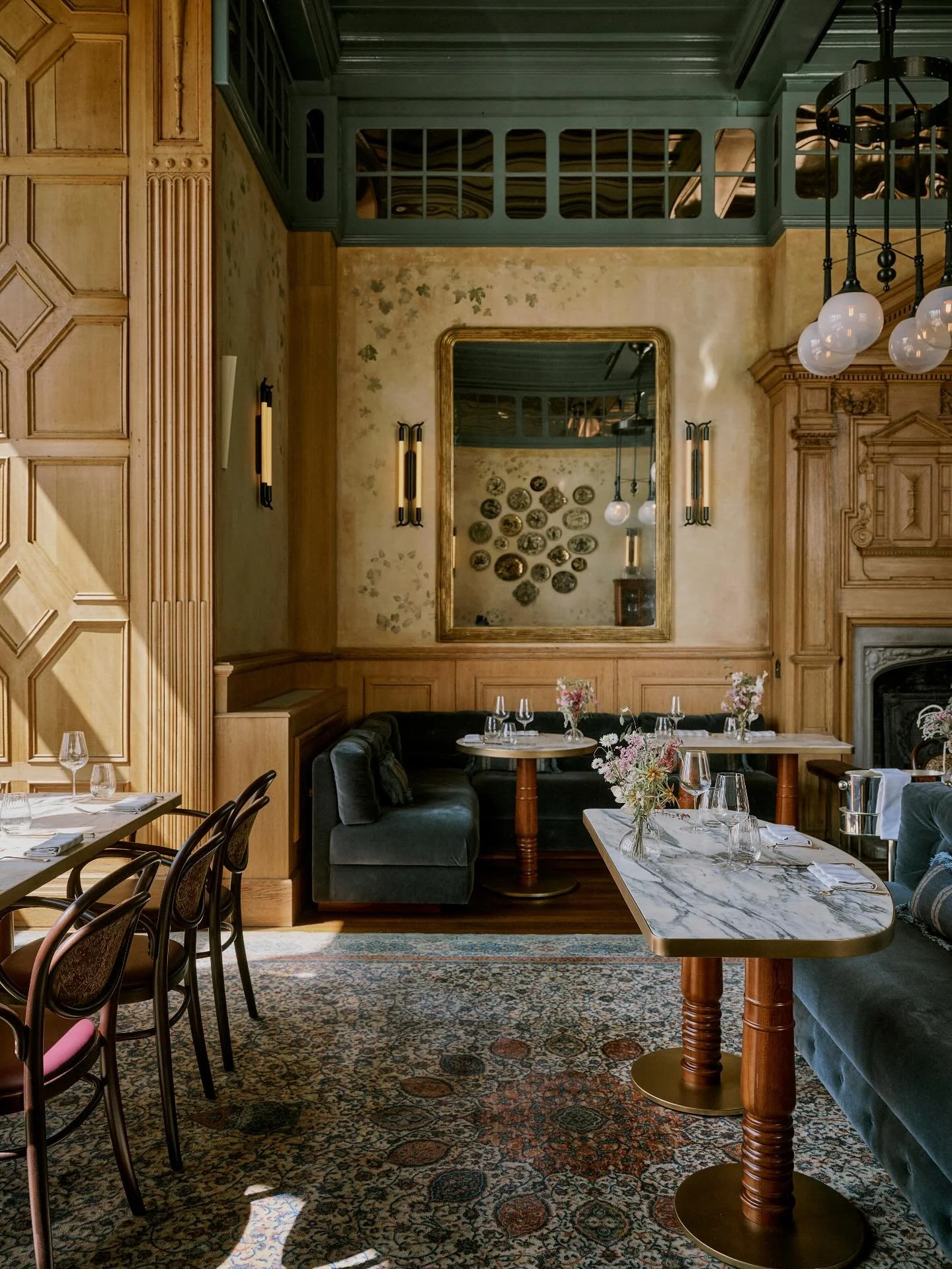 Interior of an elegant restaurant with wooden paneling, a large mirror on the wall, floral arrangements on tables, and decorative lighting fixtures.