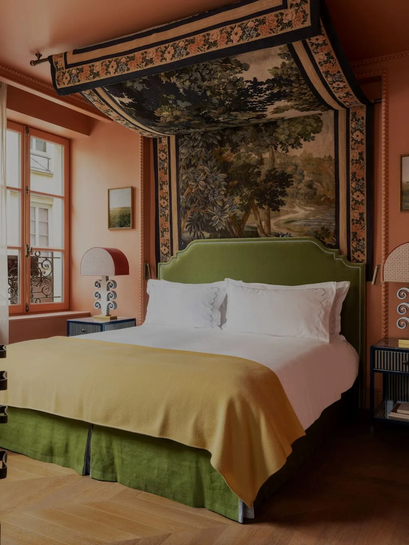 A bedroom with a bed that has a green headboard, white pillows, a yellow blanket, and a green bed skirt. There are two windows with orange frames, curtains, and a landscape painting on the wall. There are two side tables with lamps, and a large tapestry with a nature scene hanging above the bed.