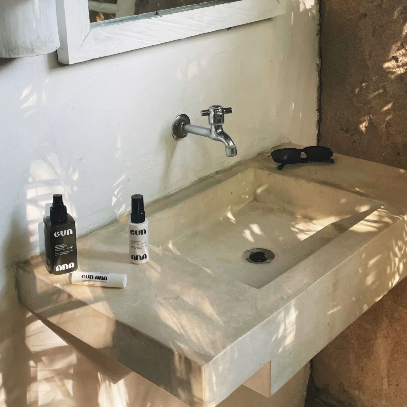 A concrete bathroom sink with a metal faucet, against an unfinished wall. On the sink are three black and white bottles and a white stick, all labeled 'CUN' and 'ANA'. To the right of the sink is a black object, possibly sunglasses.