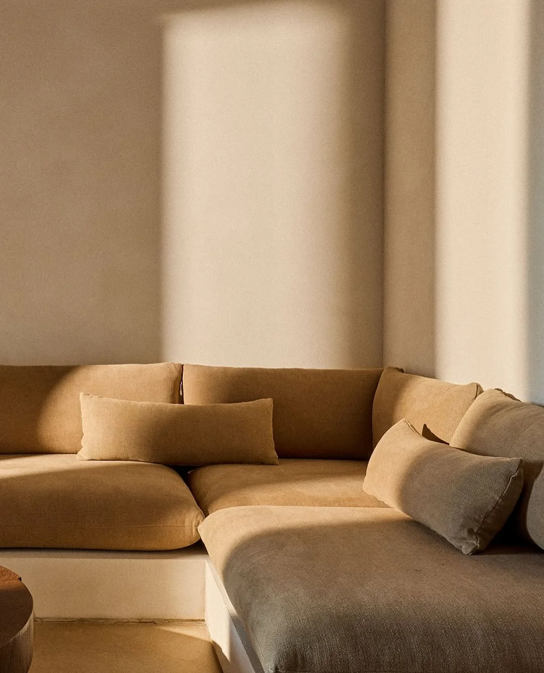 A beige sectional sofa with matching cushions in a minimally decorated room, illuminated by sunlight.