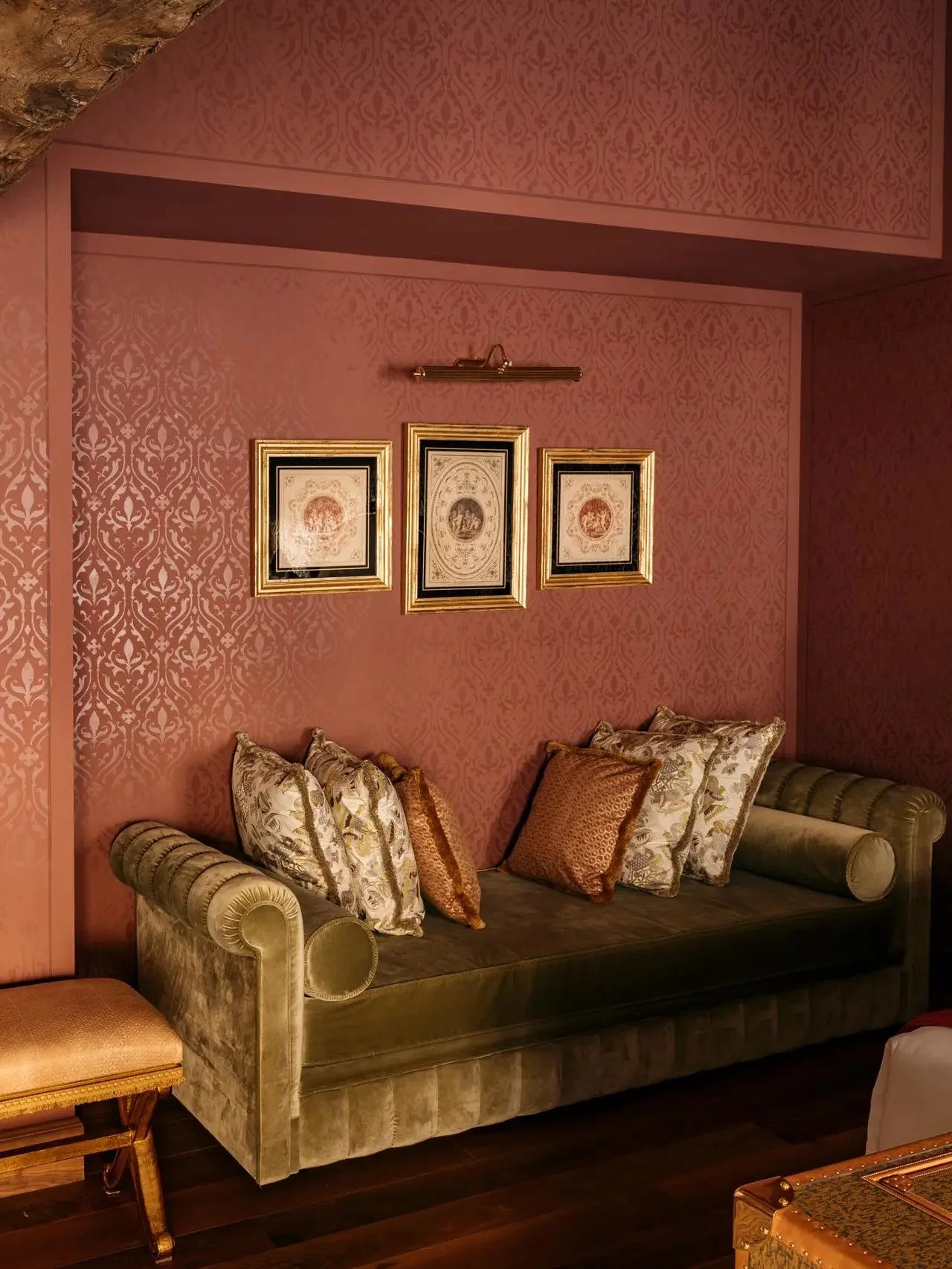 A vintage green velvet sofa with decorative pillows in gold, silver, and floral patterns, set against a pink patterned wallpapered wall with three framed art pieces and a small wall light above, in a cozy, elegant living room.