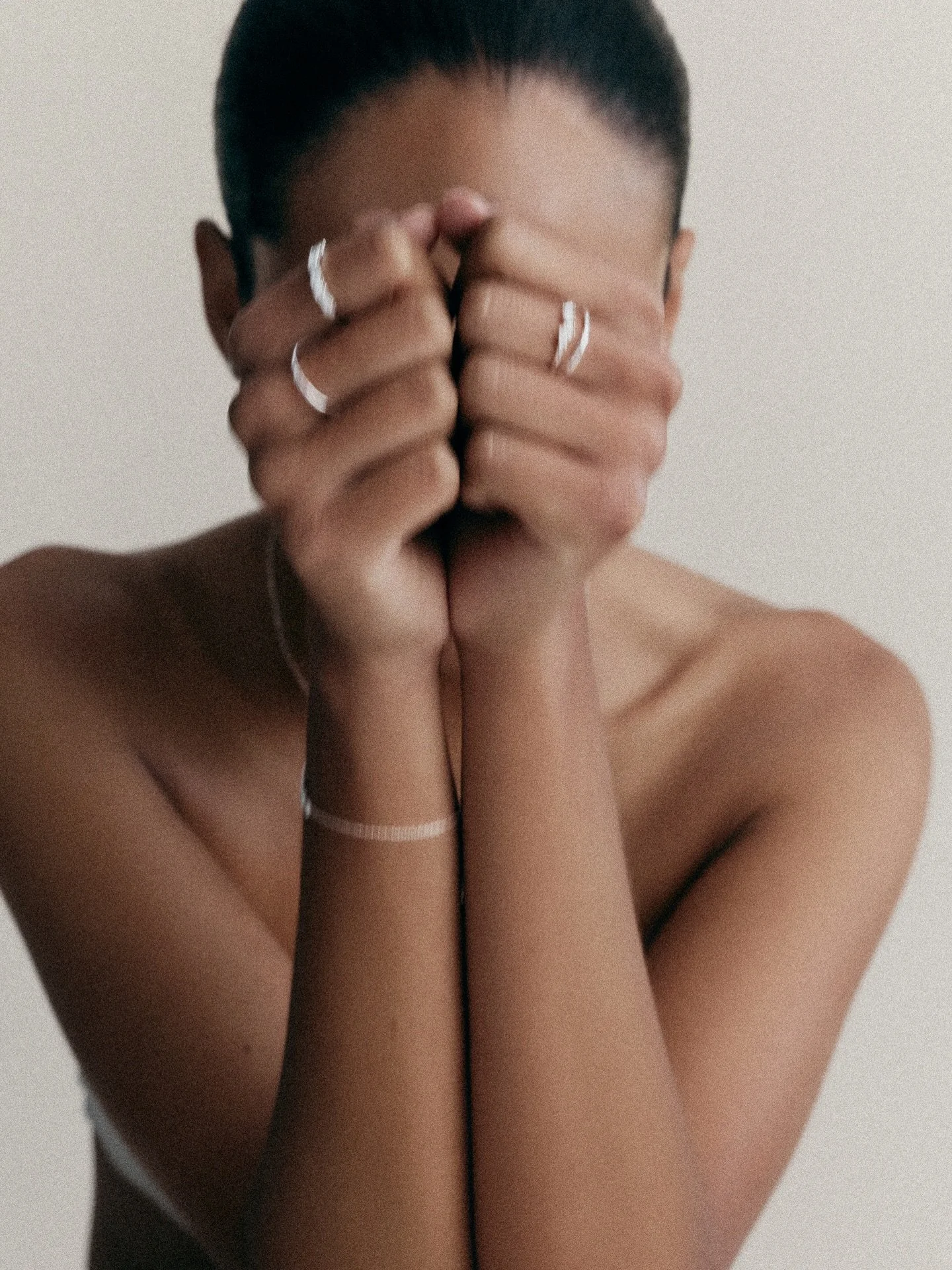 Person with short dark hair covering their face with hands, wearing rings and bracelets, against a plain neutral background.